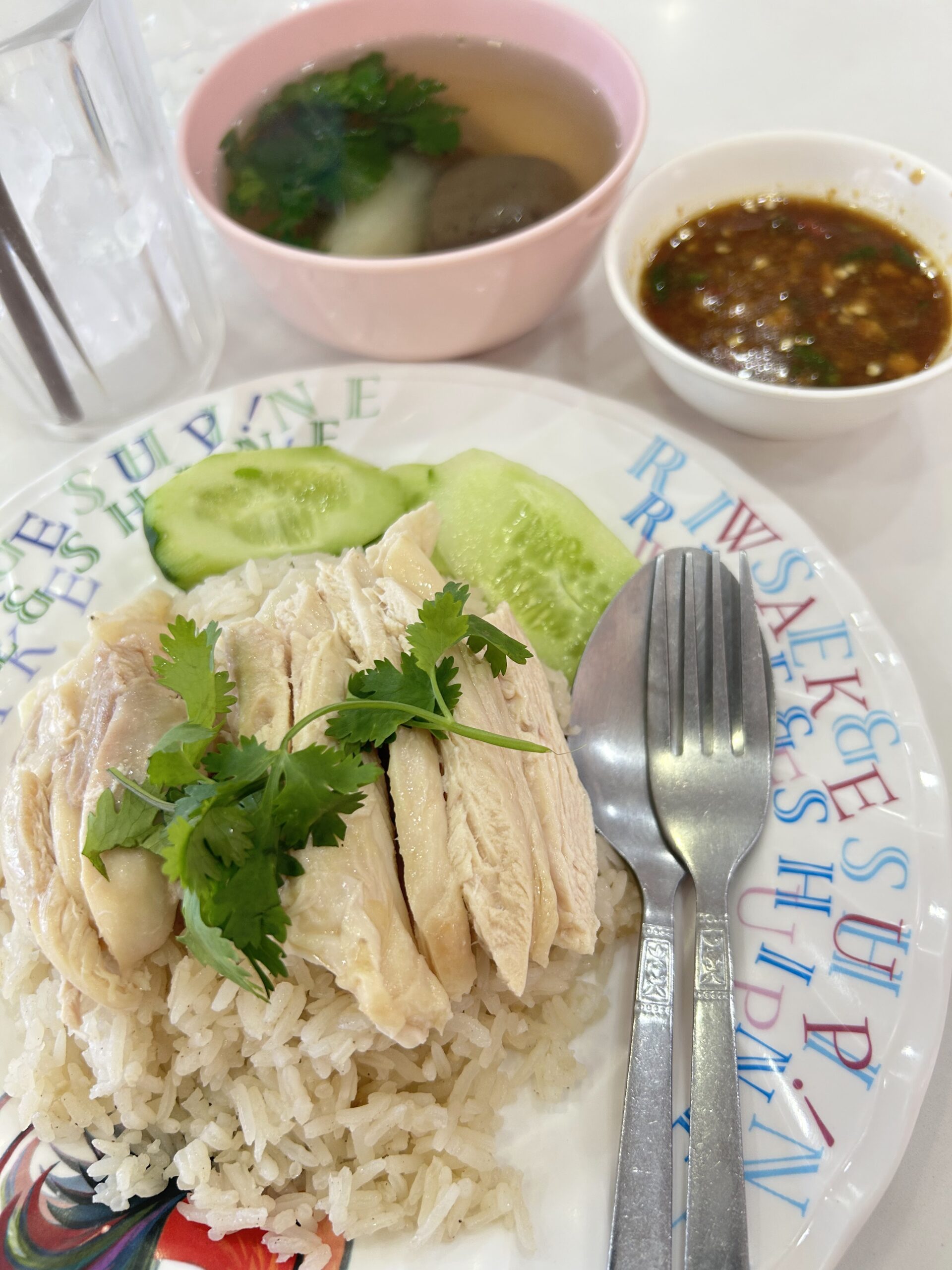 ข้าวมันไก่จานเต็มพร้อมน้ำจิ้มและน้ำซุป เสิร์ฟบนจานลายตัวอักษรสีสัน มีแตงกวาและผักชีตกแต่ง