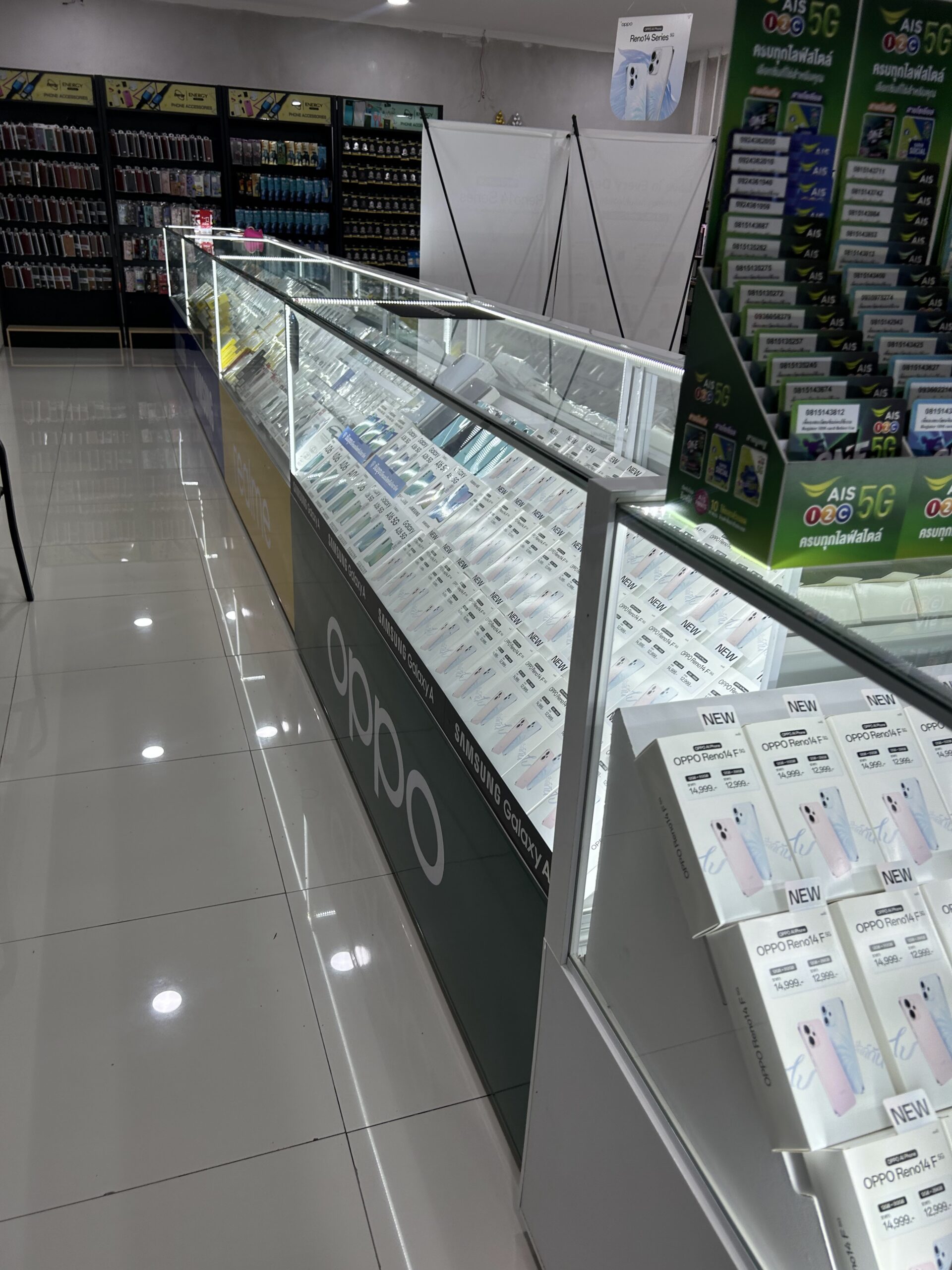ร้านขายโทรศัพท์มือถือที่มีตู้โชว์กระจกยาวแสดงมือถือหลายยี่ห้อ เช่น OPPO Samsung Galaxy และป้ายโฆษณา AIS 5G พร้อมกล่อง OPPO Reno14 F วางเรียงอยู่