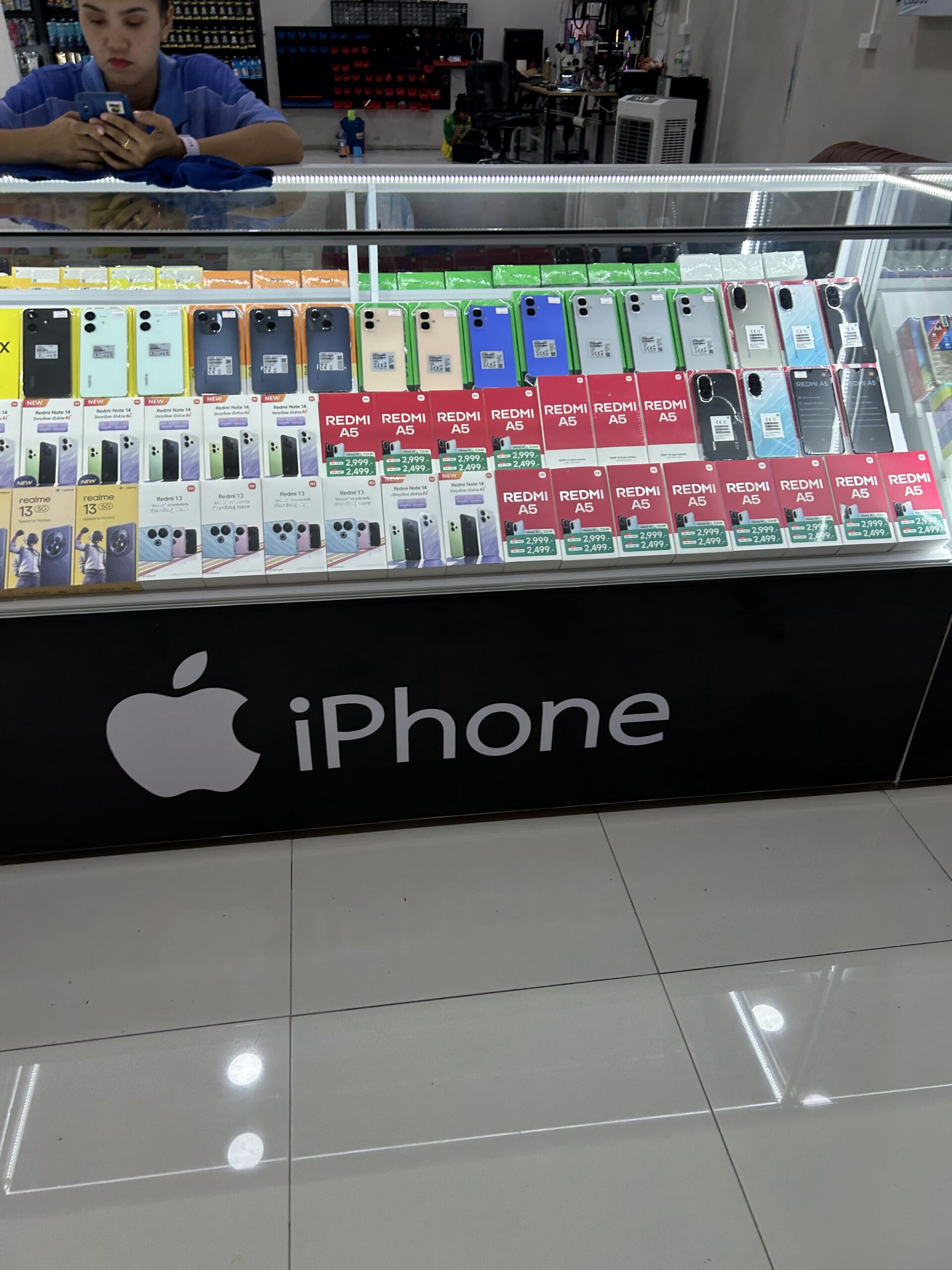 ร้านขายโทรศัพท์มือถือที่มีตู้โชว์แสดงโทรศัพท์หลายรุ่นเช่น Redmi A5 Redmi Note 14 และ Realme 13 พร้อมป้าย iPhone ของ Apple ด้านหน้าเคาน์เตอร์