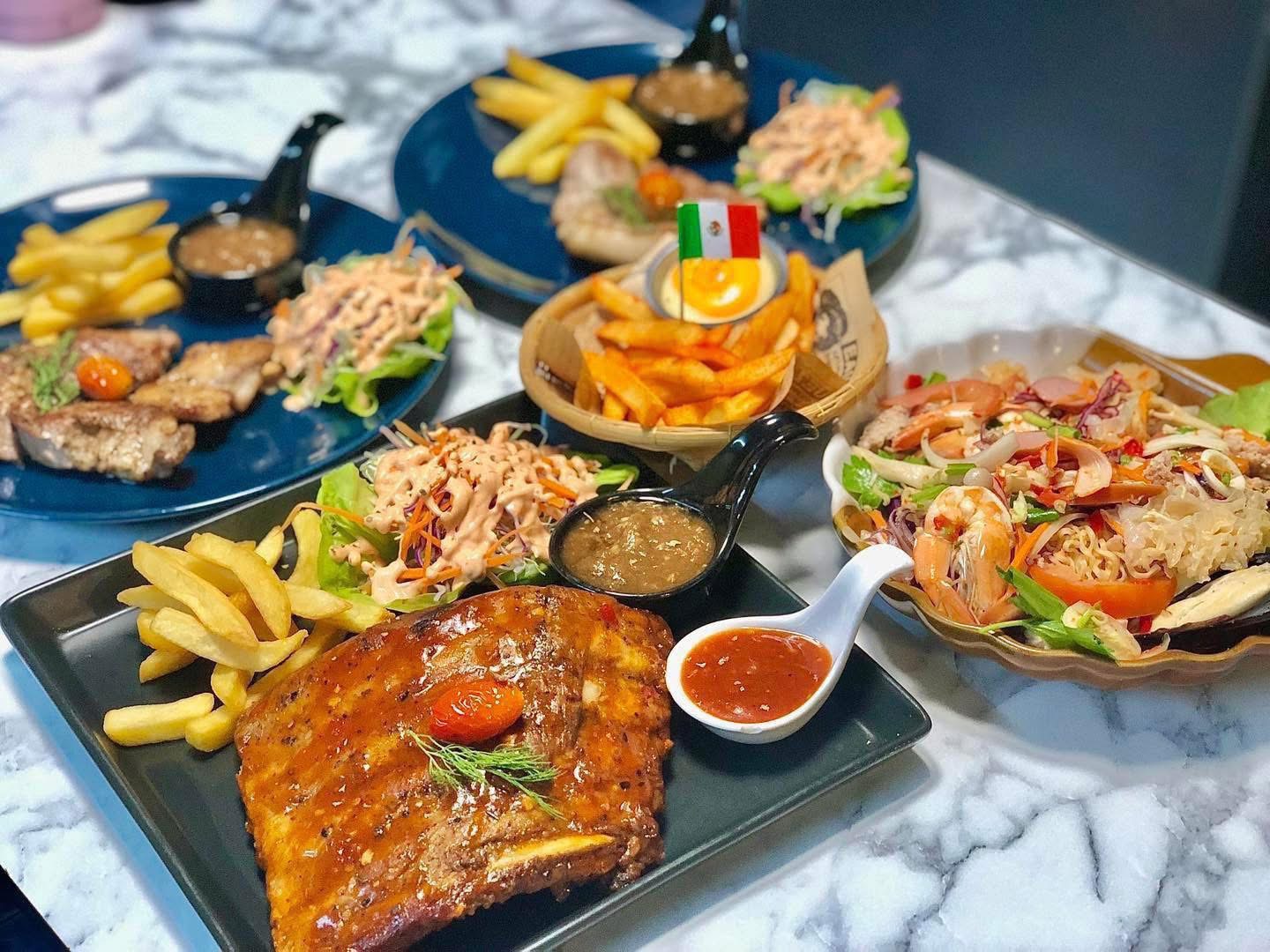 โต๊ะอาหารเต็มไปด้วยเมนูหลากหลาย ซี่โครงย่าง เฟรนช์ฟรายส์ สเต็ก สลัดผัก ยำทะเลวุ้นเส้น และเฟรนช์ฟรายส์เม็กซิกัน บนโต๊ะหินอ่อน