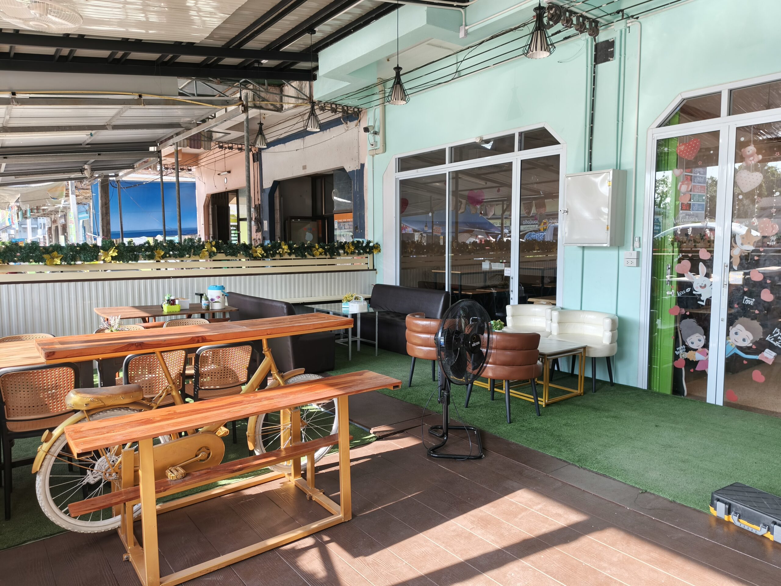 พื้นที่นั่งกลางแจ้งของร้านอาหารตกแต่งด้วยโต๊ะไม้รูปจักรยาน เก้าอี้หวาย โซฟาหนัง พัดลม และหญ้าเทียม ผนังสีเขียวมิ้นท์พร้อมประตูกระจก