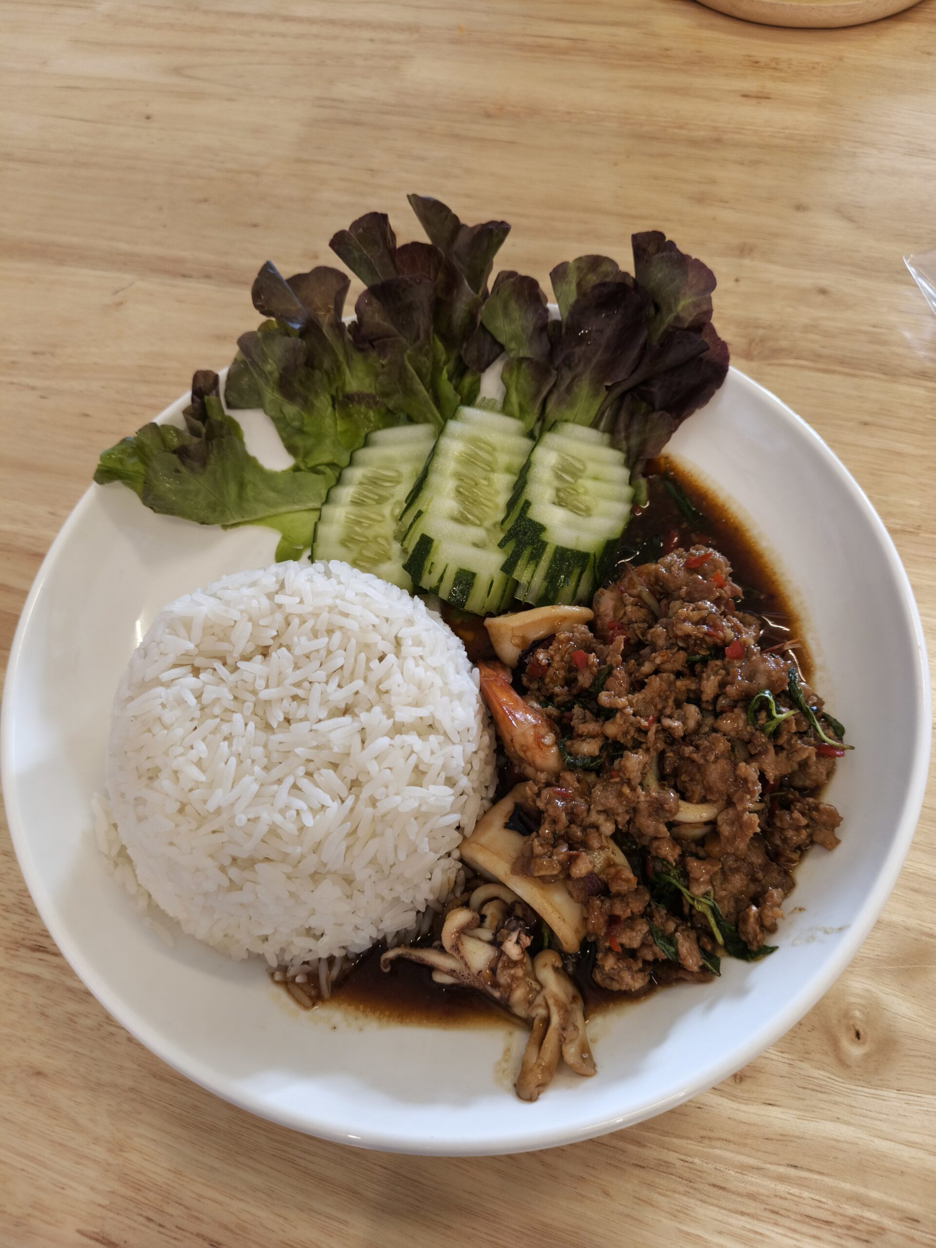 ข้าวราดกะเพราหมูสับใส่เห็ด เสิร์ฟพร้อมแตงกวาและผักสลัดบนจานขาว