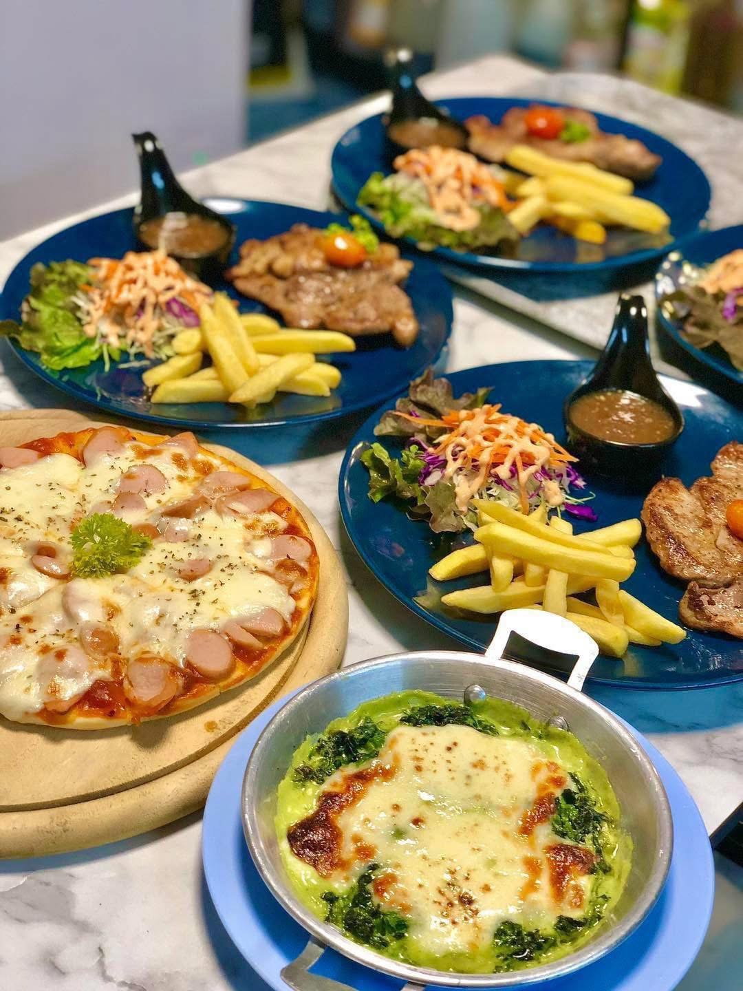 โต๊ะอาหารเต็มไปด้วยพิซซ่าหน้าไส้กรอก สเต็กเสิร์ฟพร้อมเฟรนช์ฟรายส์และสลัด และผักโขมอบชีสในจานสีฟ้า