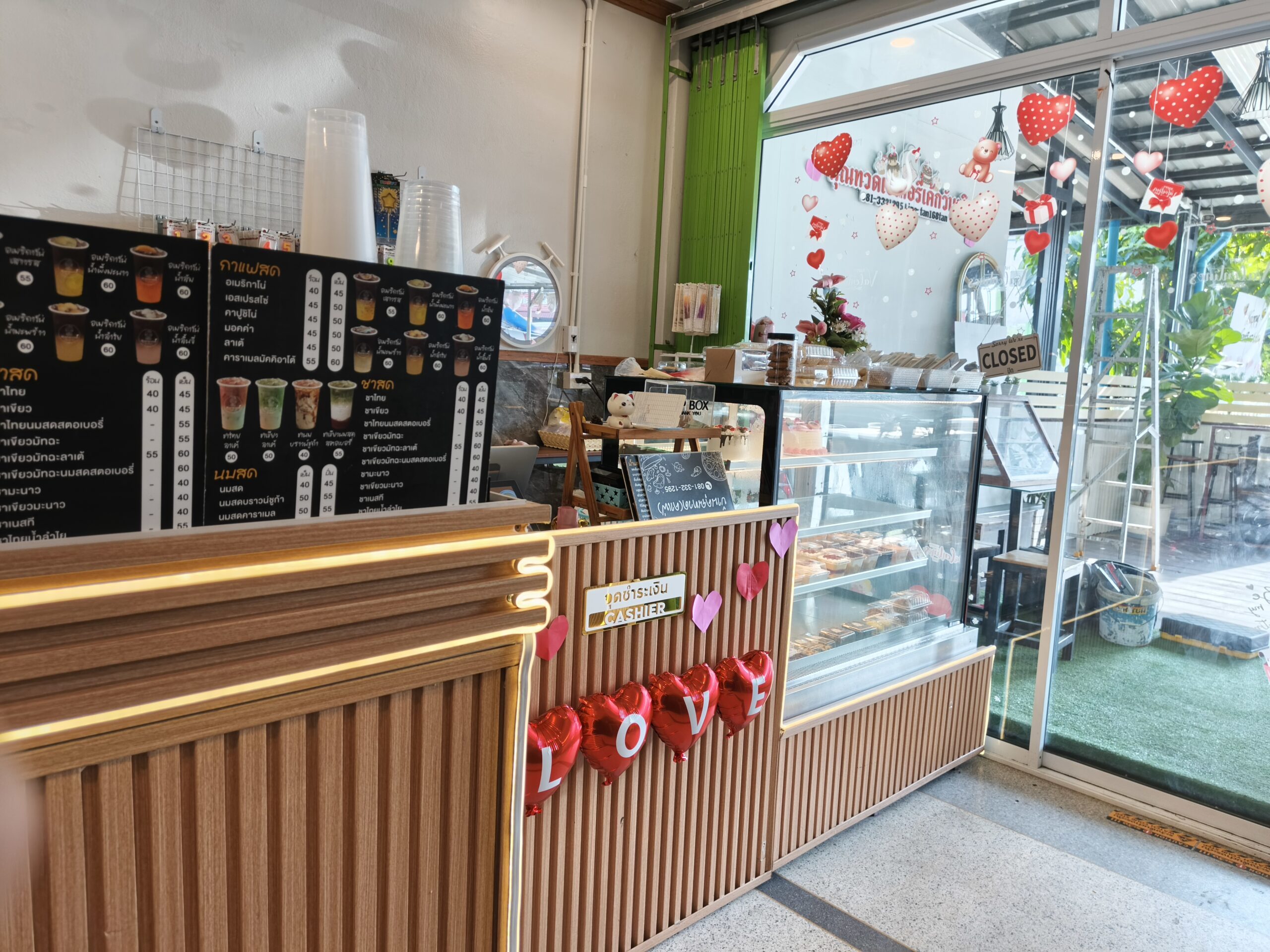 เคาน์เตอร์แคชเชียร์ร้านกาแฟตกแต่งด้วยลูกโป่งหัวใจสีแดงตัวอักษร LOVE พร้อมเมนูกาแฟสดและชาสดบนกระดานดำ มีตู้โชว์เค้กและขนม