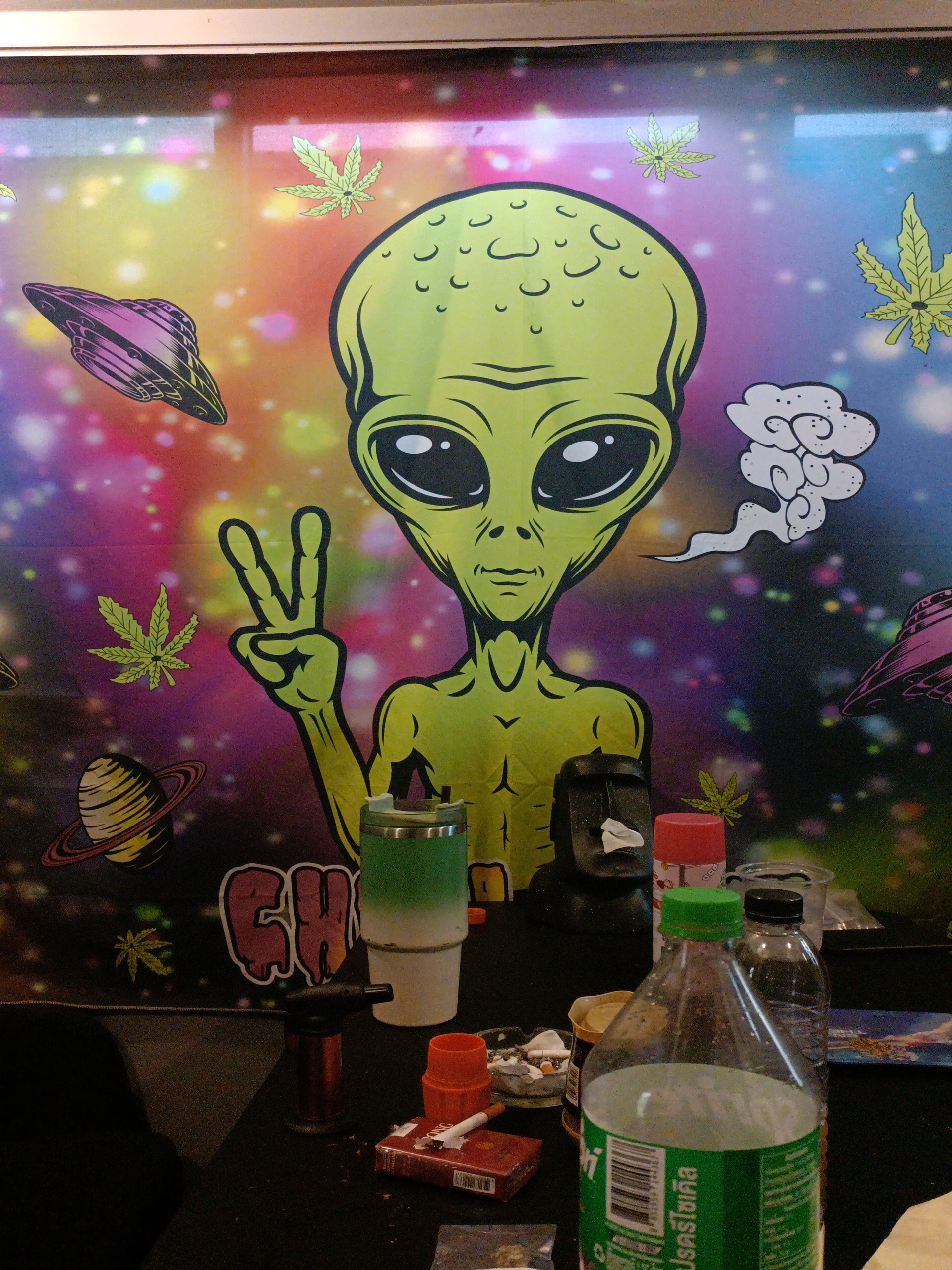 ภาพจิตรกรรมฝาผนังรูปเอเลี่ยนสีเขียวทำสัญลักษณ์สันติภาพพร้อมใบกัญชาและยานอวกาศบนพื้นหลังกาแล็กซี่สีสันสดใส