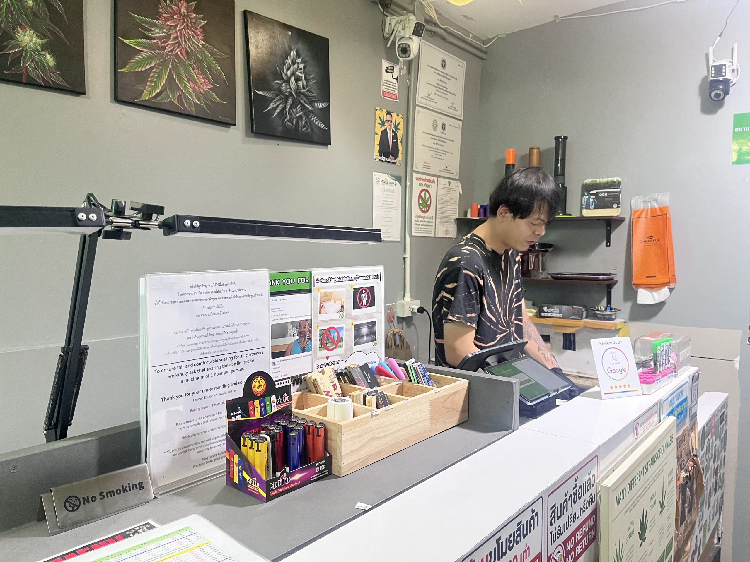 พนักงานชายยืนอยู่หลังเคาน์เตอร์ร้านกัญชา มีไฟแช็กหลากสี กระดาษมวน และป้ายห้ามสูบบุหรี่ พร้อมภาพวาดใบกัญชาบนผนัง