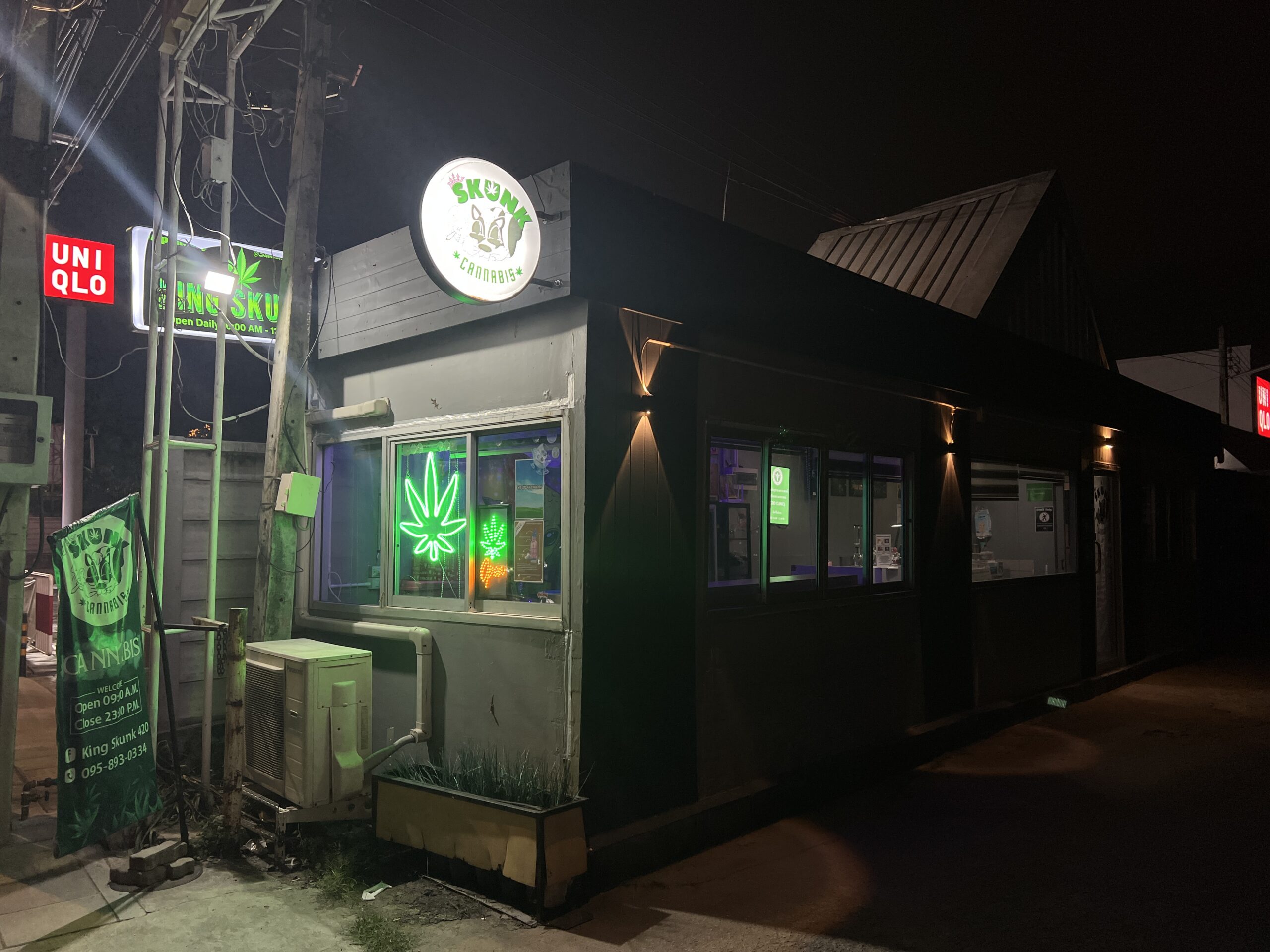 ร้าน King Skunk Cannabis ถ่ายตอนกลางคืน มีป้ายไฟนีออนรูปใบกัญชาสีเขียวและป้ายวงกลมเรืองแสง ตั้งอยู่ใกล้ร้าน Uniqlo