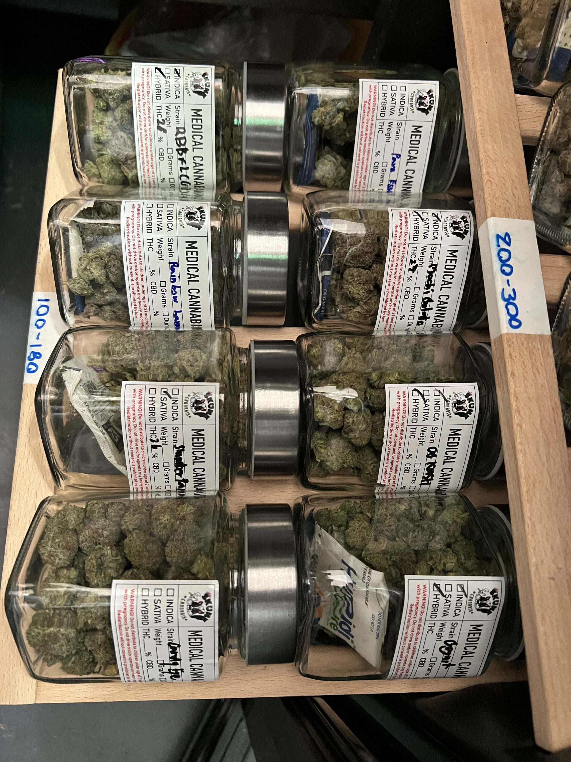 ขวดโหลแก้วบรรจุกัญชาทางการแพทย์หลายสายพันธุ์วางเรียงบนชั้นไม้พร้อมฉลากระบุชื่อสายพันธุ์