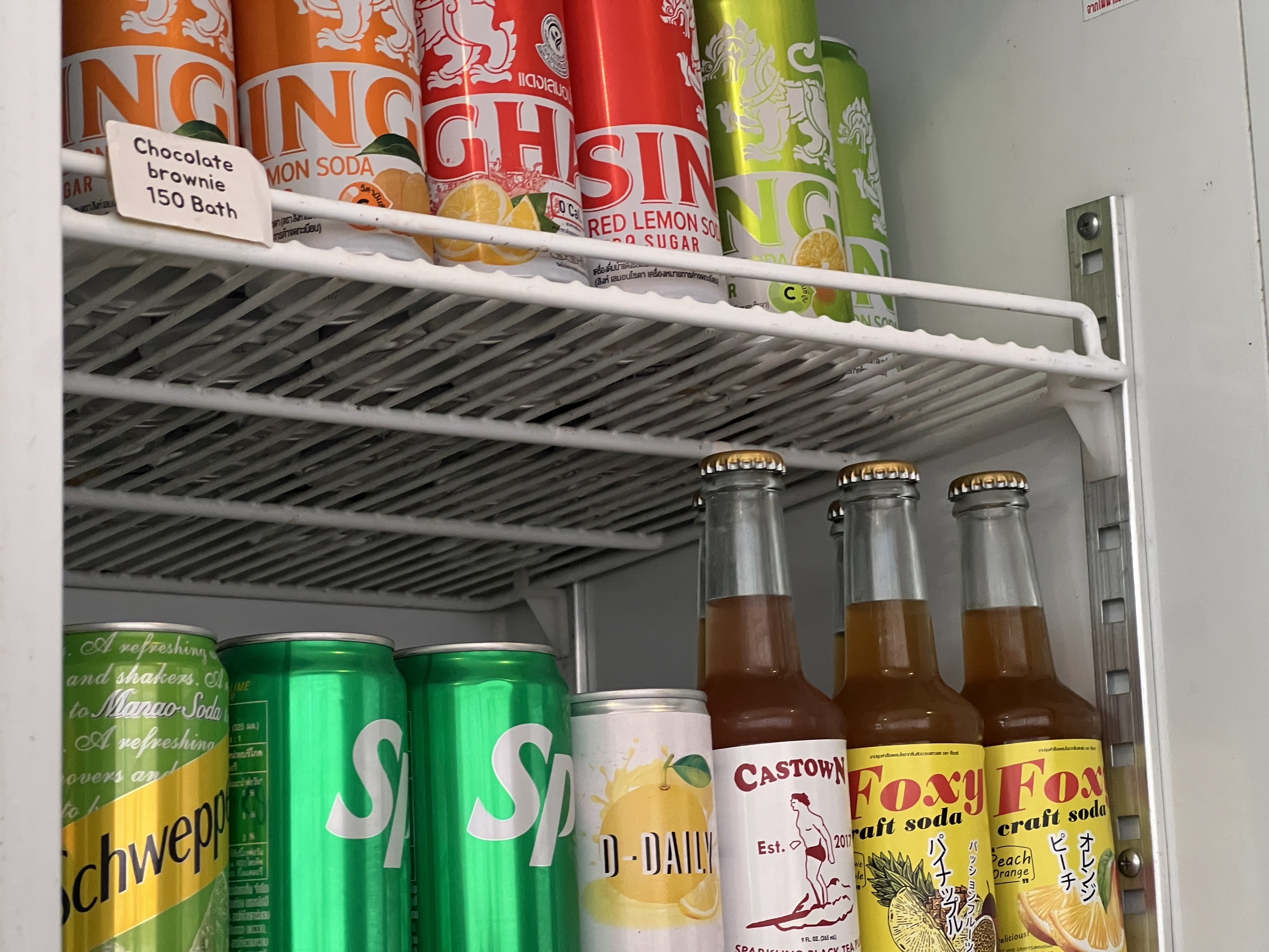 ตู้เย็นแสดงเครื่องดื่มหลากหลายชนิด ได้แก่ น้ำอัดลมสิงห์ ชเวปส์ สไปรท์ โซดา Foxy craft soda และ Castown พร้อมป้ายราคาช็อกโกแลตบราวนี่ 150 บาท