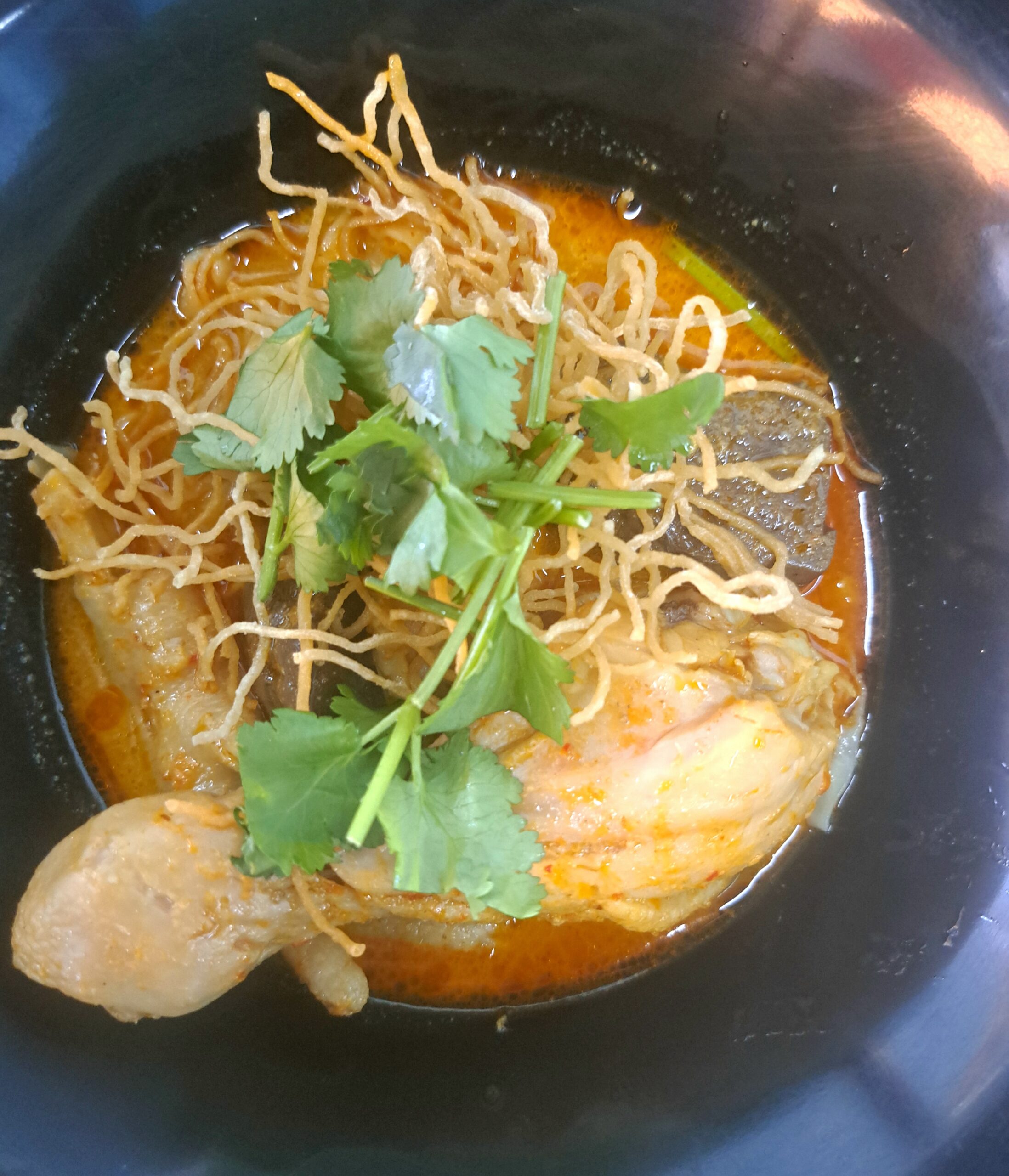 ข้าวซอยไก่ในชามดำ น้ำซุปแกงกะหรี่สีส้มทอง โรยบะหมี่กรอบและผักชี มีน่องไก่ชิ้นใหญ่