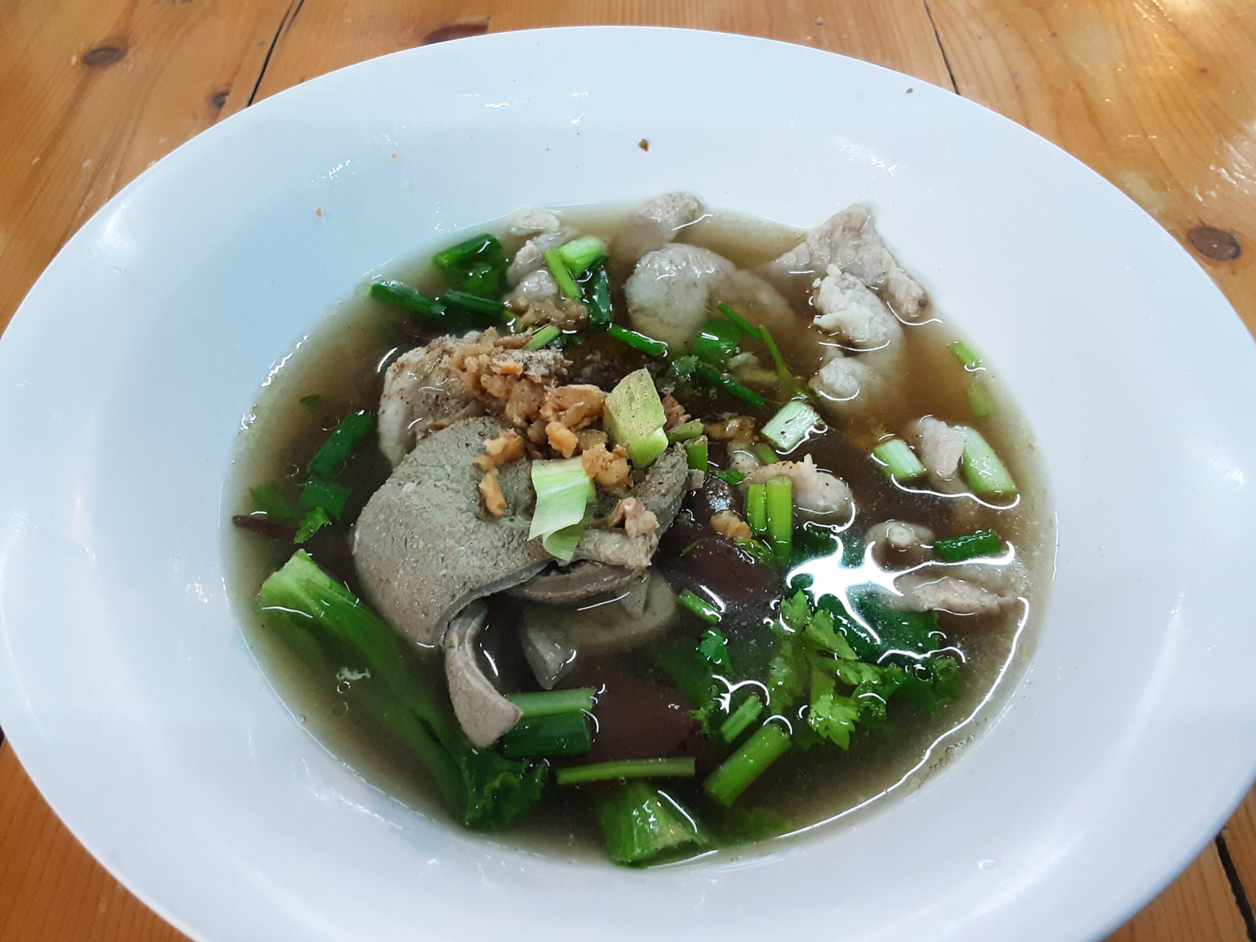 ก๋วยเตี๋ยวเครื่องในหมูน้ำใส ในชามสีขาว มีตับ ลูกชิ้น เลือดหมู ผักชี และต้นหอม บนโต๊ะไม้