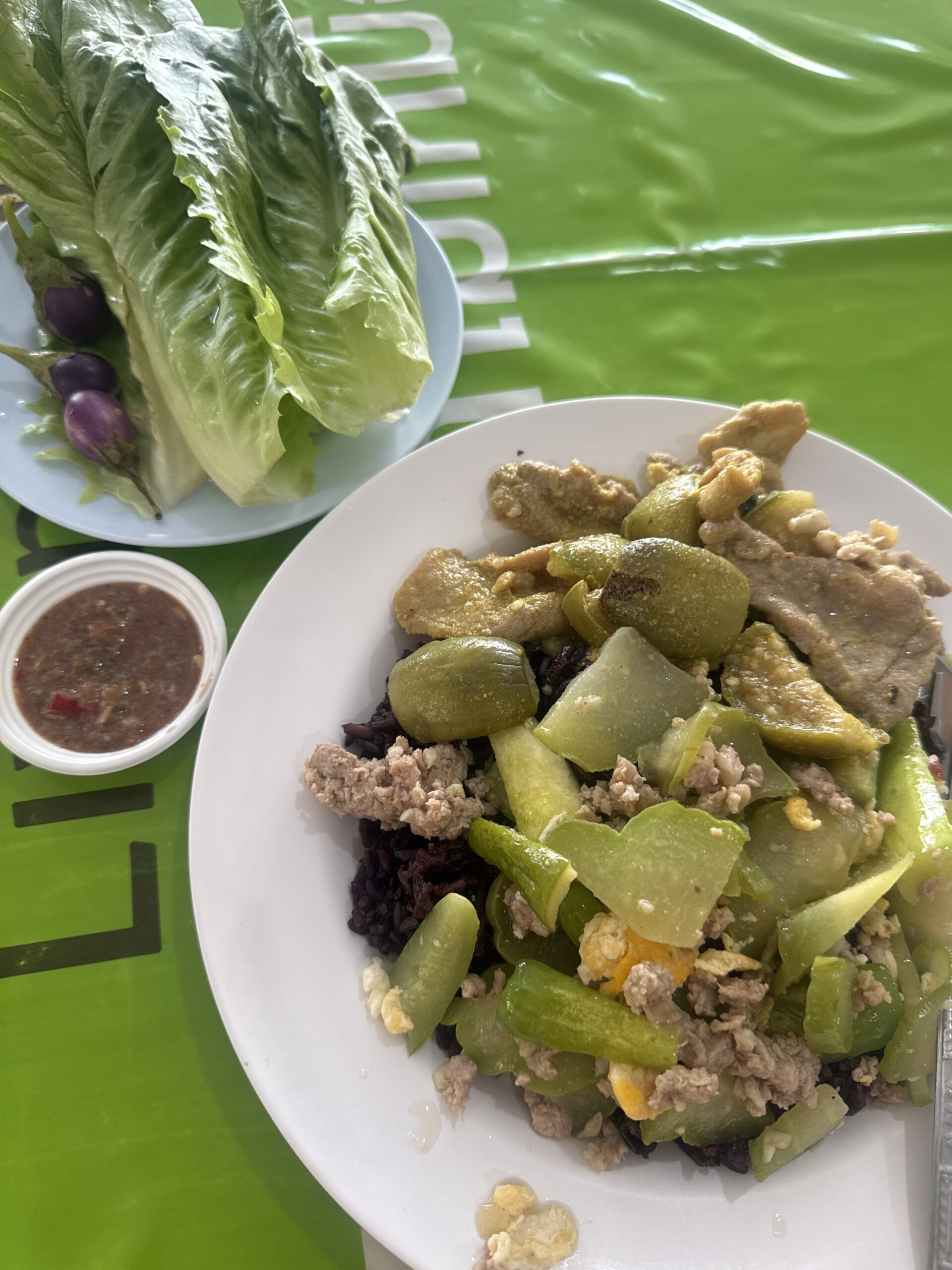 จานข้าวไรซ์เบอร์รี่ราดผัดบวบใส่หมูสับและไข่ พร้อมผักสดและน้ำพริกกะปิบนโต๊ะสีเขียว