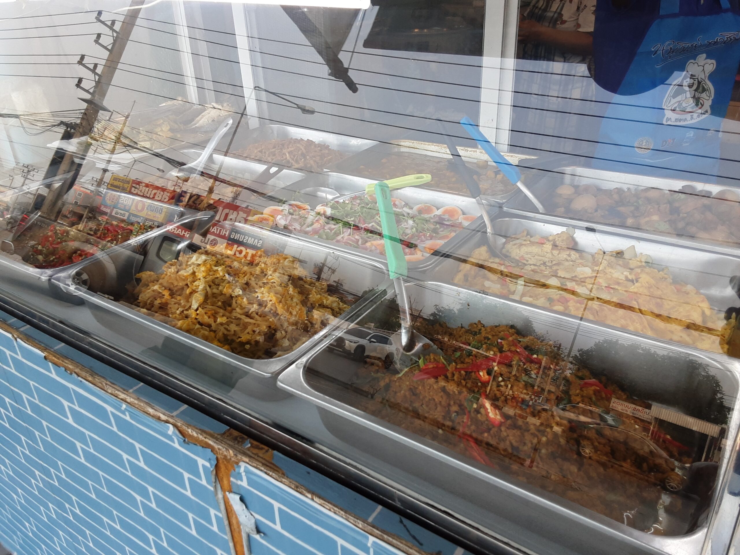 ตู้โชว์อาหารตามสั่งริมถนน มีกับข้าวหลายอย่างในถาดสแตนเลส เช่น ผัดกะเพรา แกงส้ม ยำ และไข่พะโล้ มีกระจกกันฝุ่นด้านบน