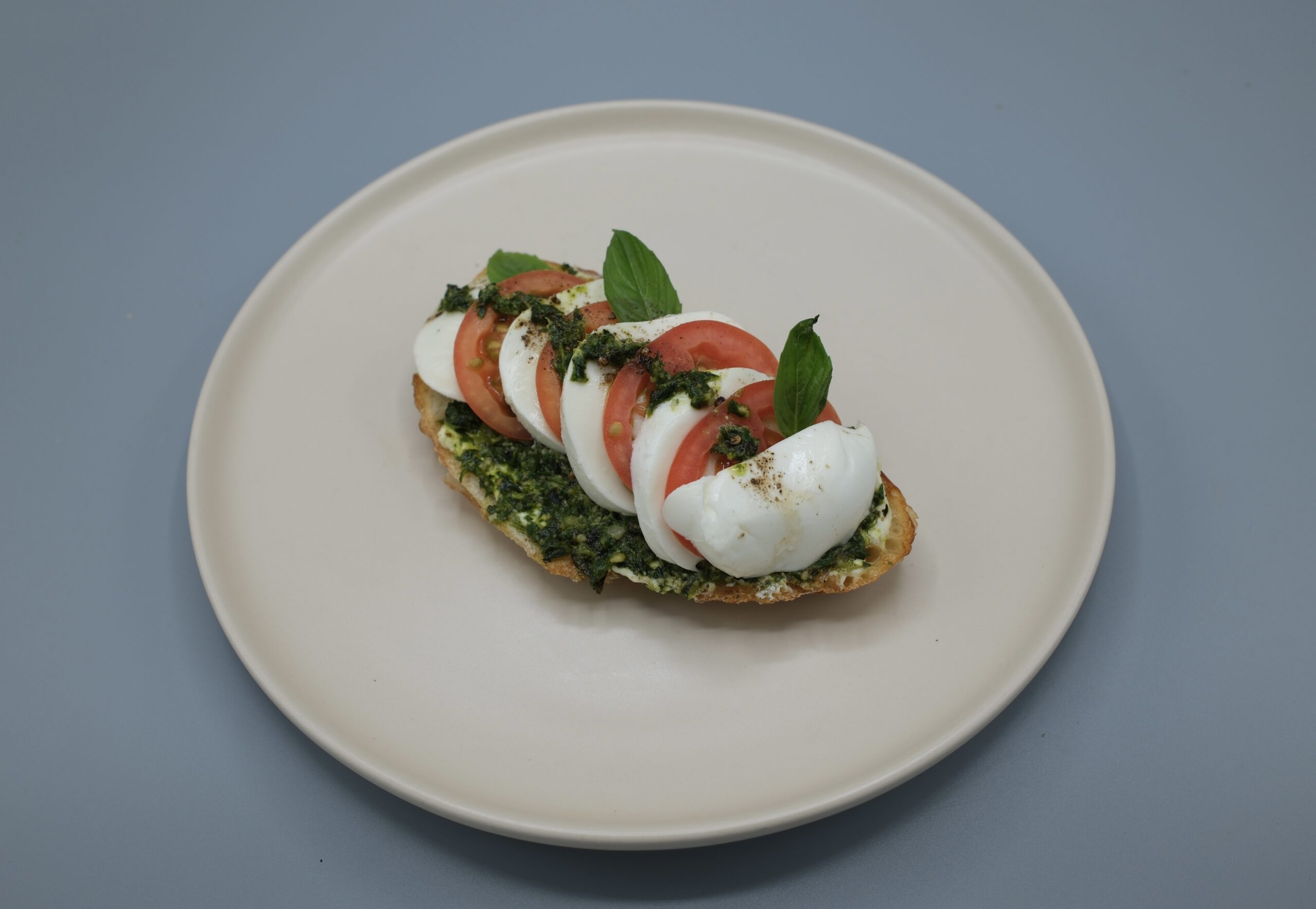 บรูสเก็ตต้าหน้ามอสซาเรลล่าชีสกับมะเขือเทศสด ราดซอสเพสโต้ ตกแต่งด้วยใบโหระพาอิตาเลียน บนจานสีขาว