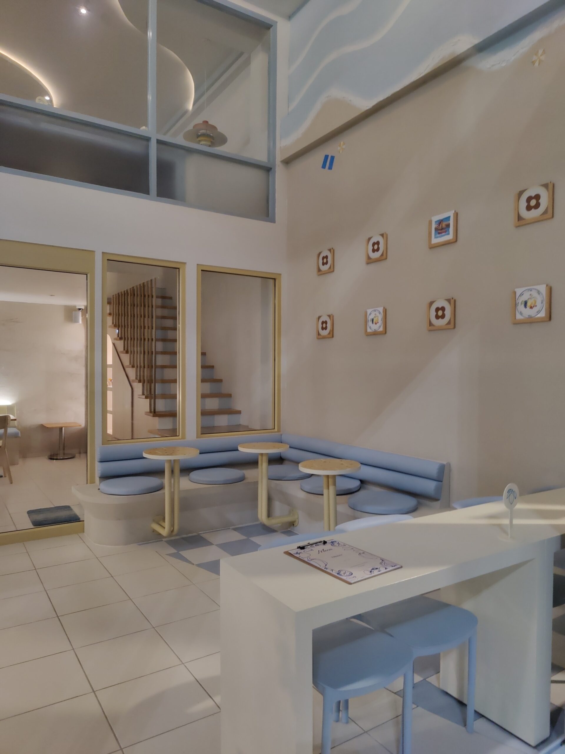 ภายในร้านคาเฟ่โทนสีฟ้าพาสเทลและสีขาว มีโซฟายาวสีฟ้า โต๊ะกลมไม้ กรอบรูปตกแต่งผนัง และบันไดไม้ บรรยากาศน่ารักสไตล์กรีก