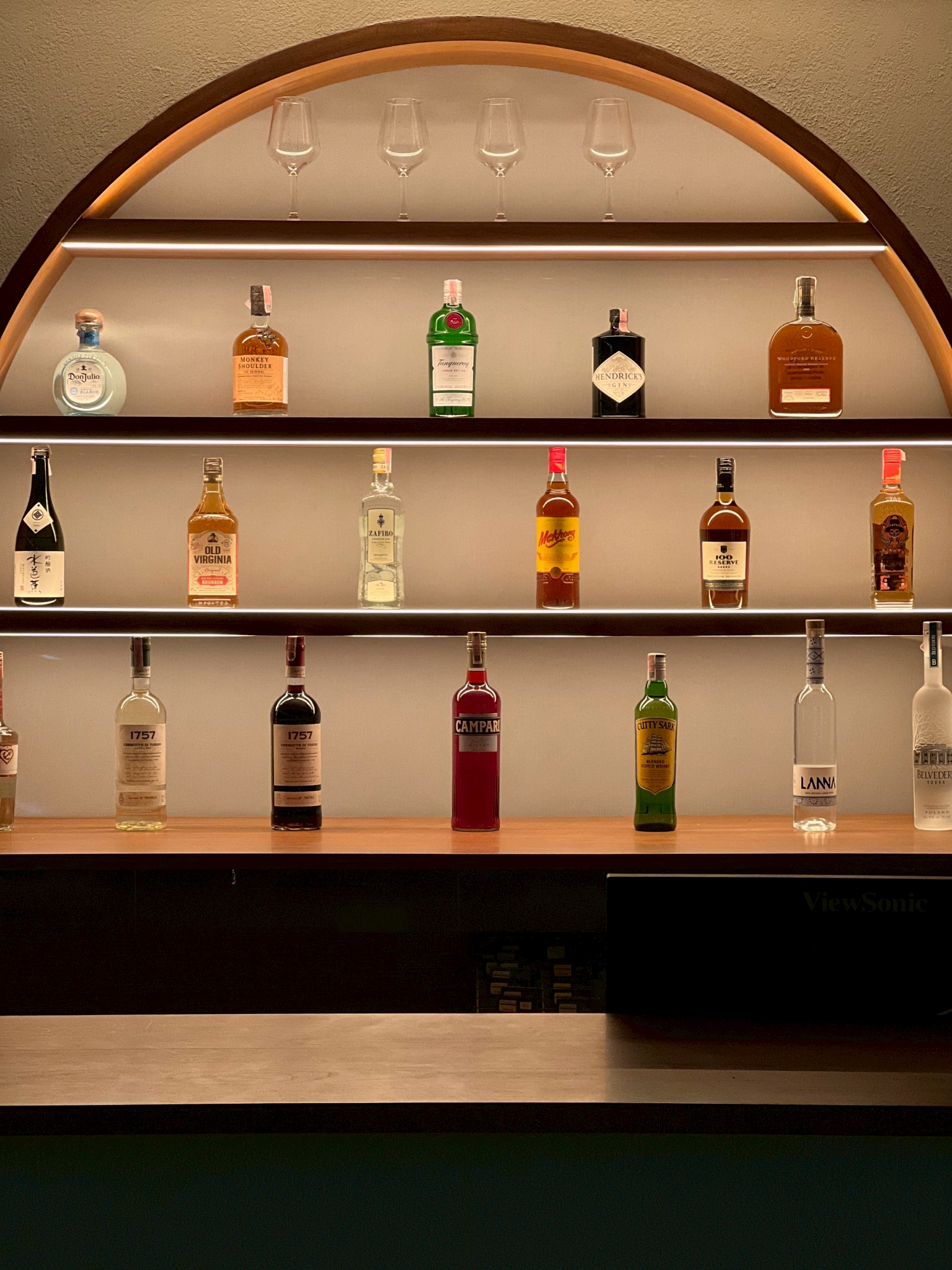 ชั้นวางเครื่องดื่มแอลกอฮอล์หลากหลายยี่ห้อในตู้โค้งมีไฟส่องสว่าง พร้อมแก้วไวน์เรียงบนชั้นบนสุด