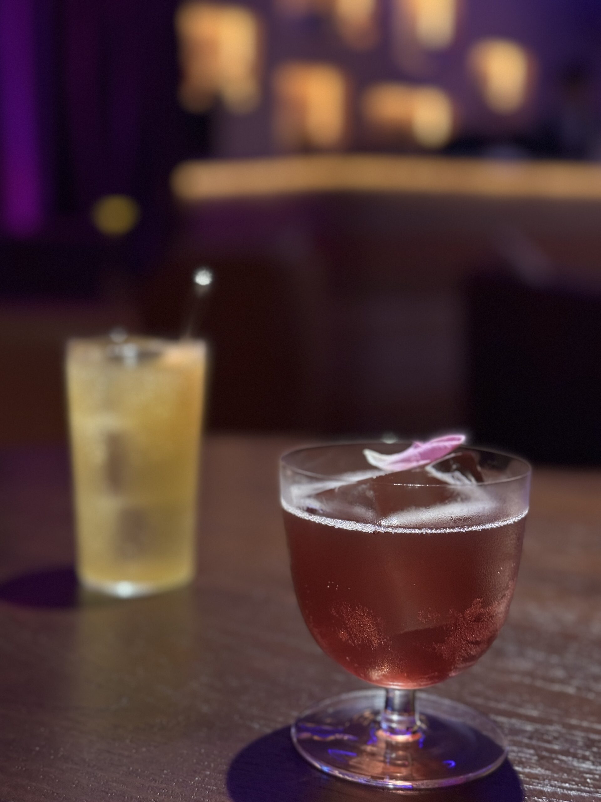 ค็อกเทลสีแดงตกแต่งด้วยกลีบดอกไม้และเครื่องดื่มสีเหลืองในแก้วทรงสูงบนโต๊ะบาร์บรรยากาศแสงสีม่วง