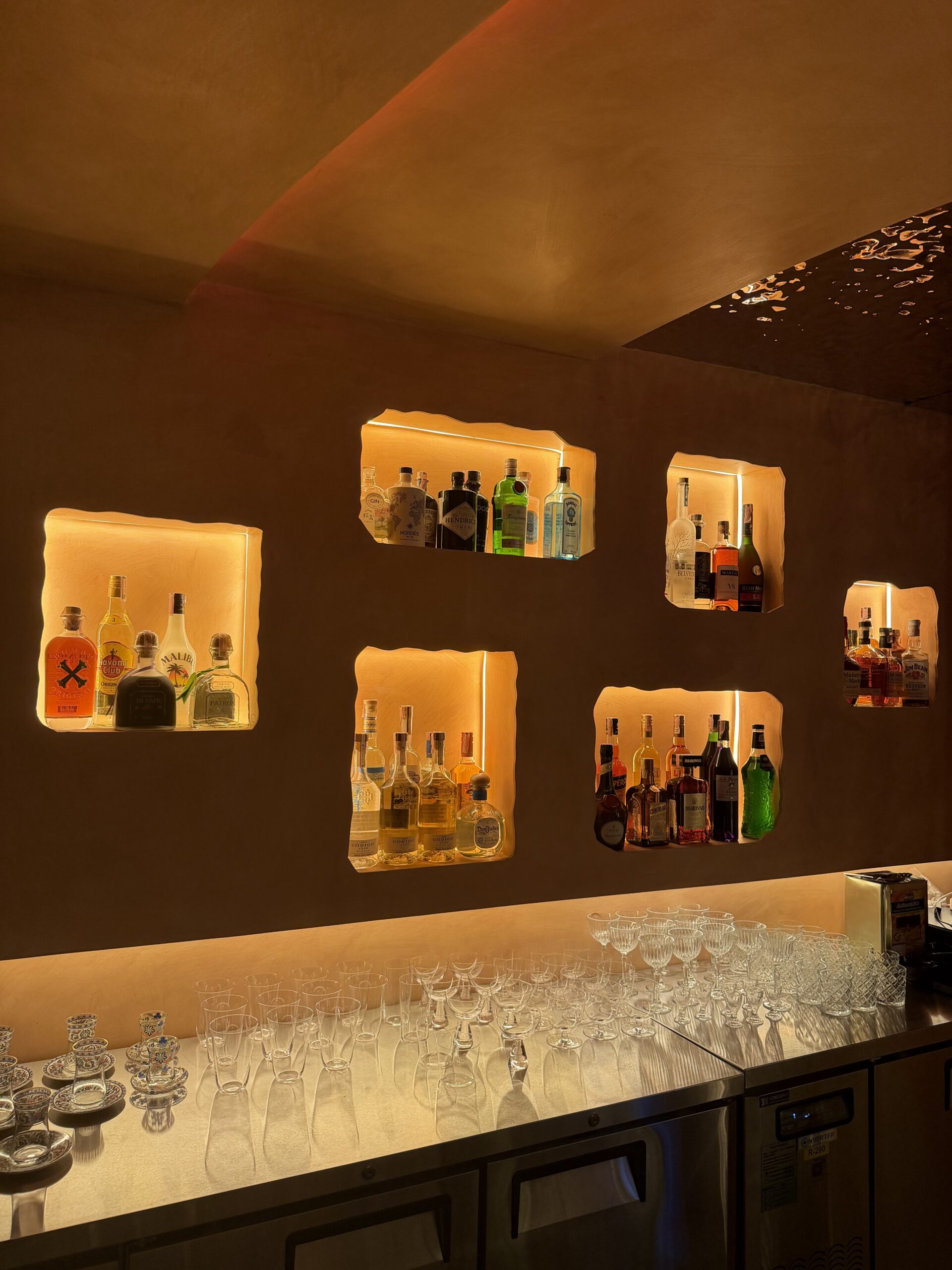 บาร์เครื่องดื่มที่มีช่องเก็บขวดเหล้าฝังผนังพร้อมไฟส่องสว่างสีอบอุ่น แก้วค็อกเทลหลากหลายแบบเรียงบนเคาน์เตอร์
