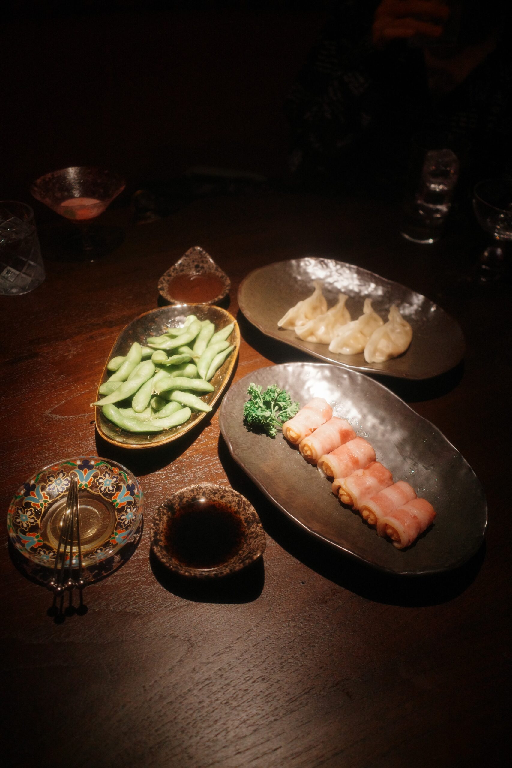 จานอาหารญี่ปุ่นสไตล์อิซากายะบนโต๊ะไม้ในบรรยากาศสลัว มีถั่วแระญี่ปุ่น เกี๊ยวซ่านึ่ง เบคอนม้วน พร้อมซอสจิ้มและเครื่องดื่ม