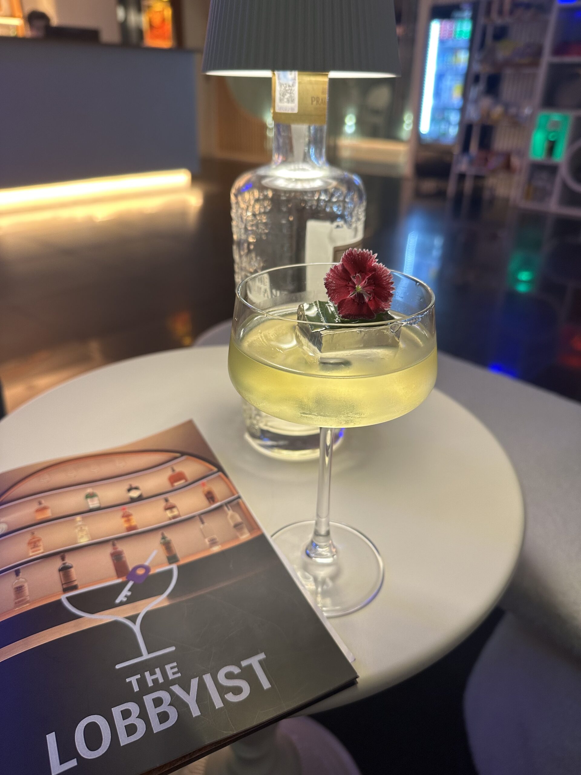 ค็อกเทลสีเหลืองในแก้วคูเป้ตกแต่งด้วยดอกไม้สีแดง วางบนโต๊ะกลมสีขาวข้างเมนูร้าน The Lobbyist
