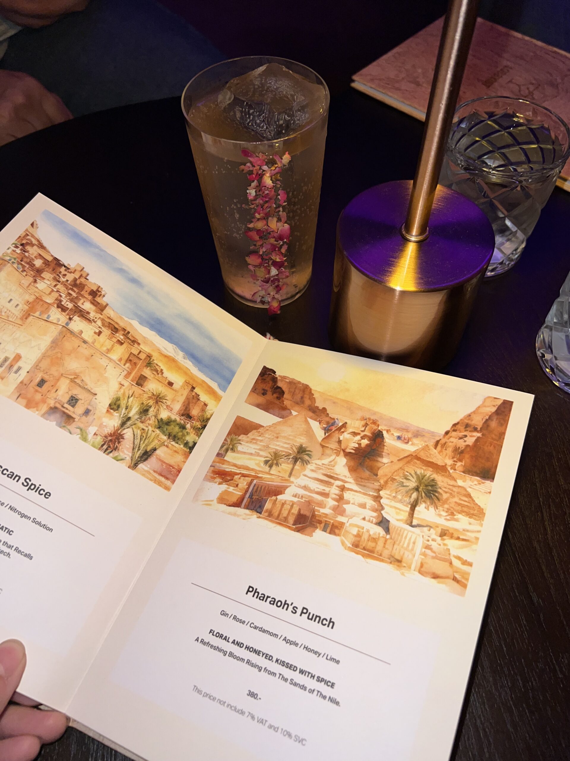 เมนูค็อกเทล Pharaoh's Punch พร้อมเครื่องดื่มที่ตกแต่งด้วยกลีบกุหลาบในแก้วใส ส่วนผสมประกอบด้วยจิน กุหลาบ กระวาน แอปเปิ้ล น้ำผึ้ง และมะนาว ราคา 380 บาท