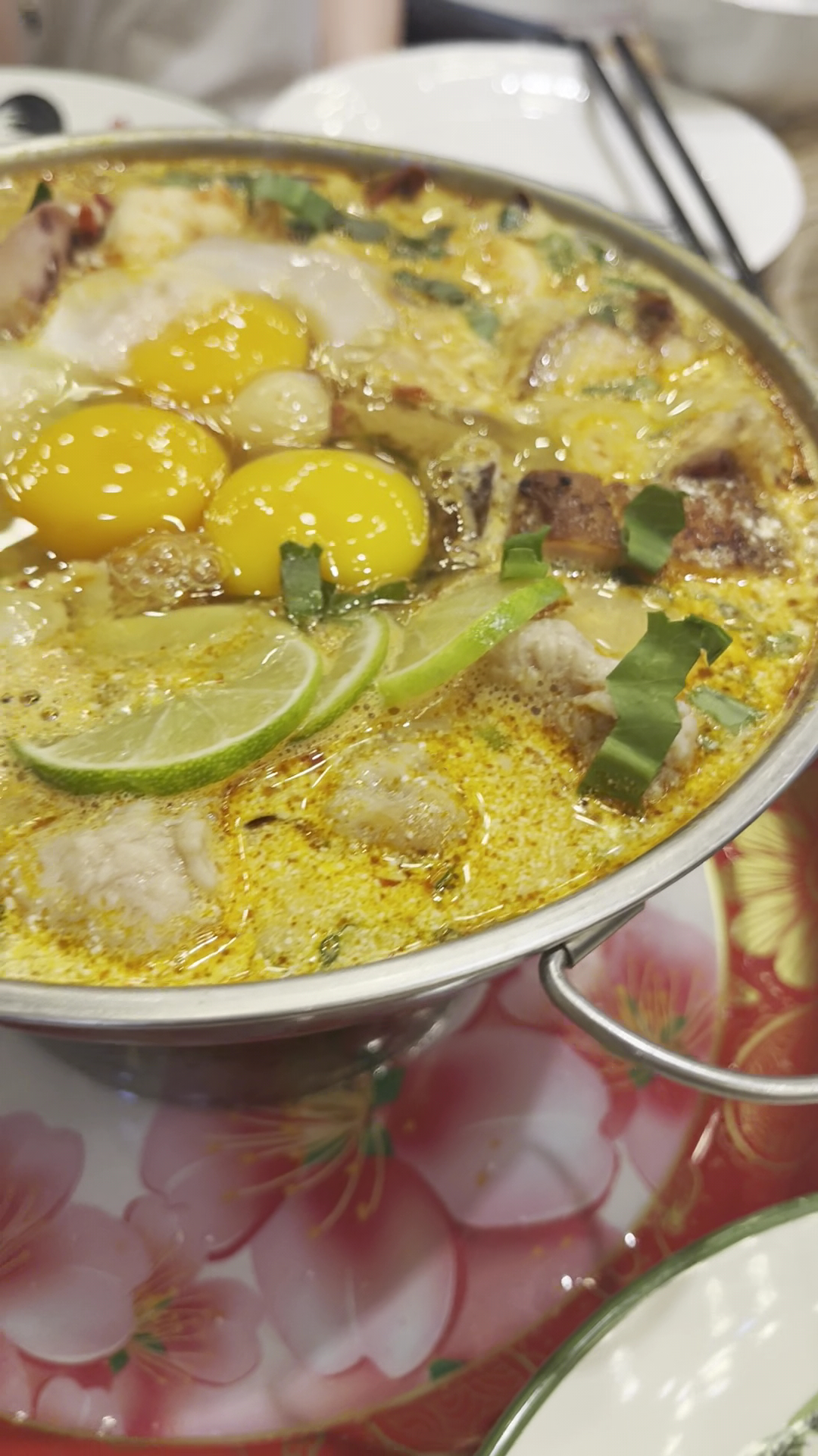 หม้อไฟอาหารทะเลในน้ำซุปสีเหลืองทองใส่ไข่แดง มะนาว ผักชี และเนื้อปลา เสิร์ฟในหม้อสแตนเลสบนโต๊ะลายดอกไม้