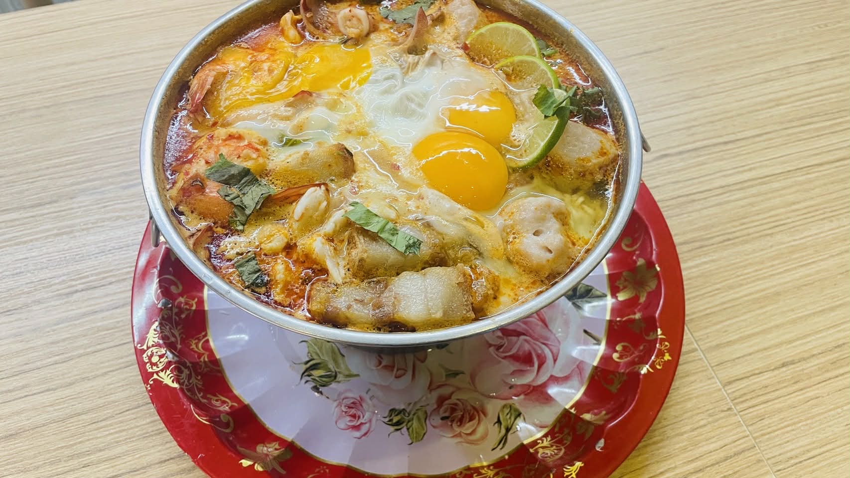 หม้อต้มยำทะเลใส่ไข่ดาวสองฟองพร้อมกุ้ง หมึก และมะนาว เสิร์ฟในหม้อสแตนเลสบนจานลายดอกกุหลาบ