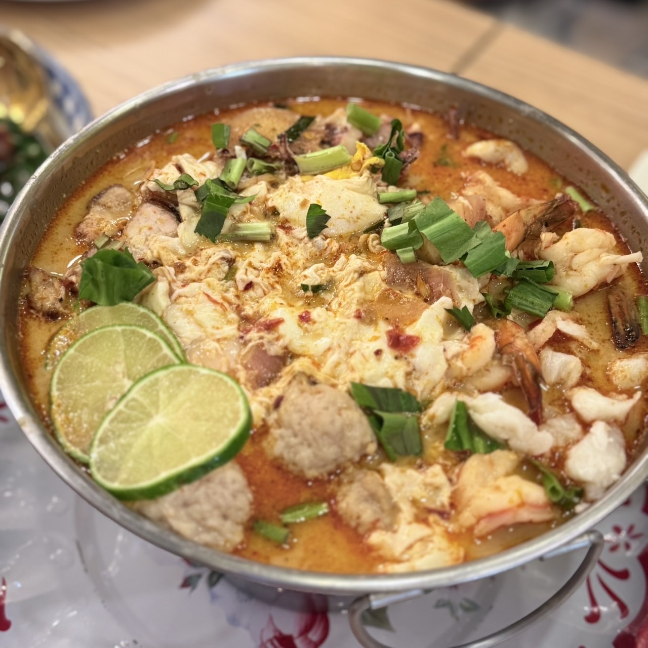 ต้มยำทะเลรวมมิตรในหม้อสเตนเลส มีกุ้ง ลูกชิ้น ไข่ และมะนาวฝานเป็นแว่น โรยต้นหอมสด น้ำซุปสีส้มแดงเข้มข้น