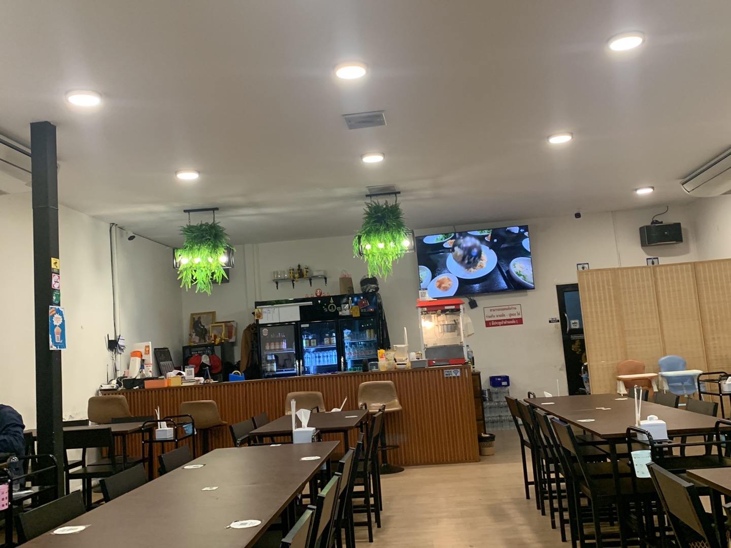 บรรยากาศภายในร้านอาหารไทย มีโต๊ะเก้าอี้สีเข้ม เคาน์เตอร์บาร์ ตู้เย็นเครื่องดื่ม จอทีวีขนาดใหญ่ และต้นไม้แขวนตกแต่ง
