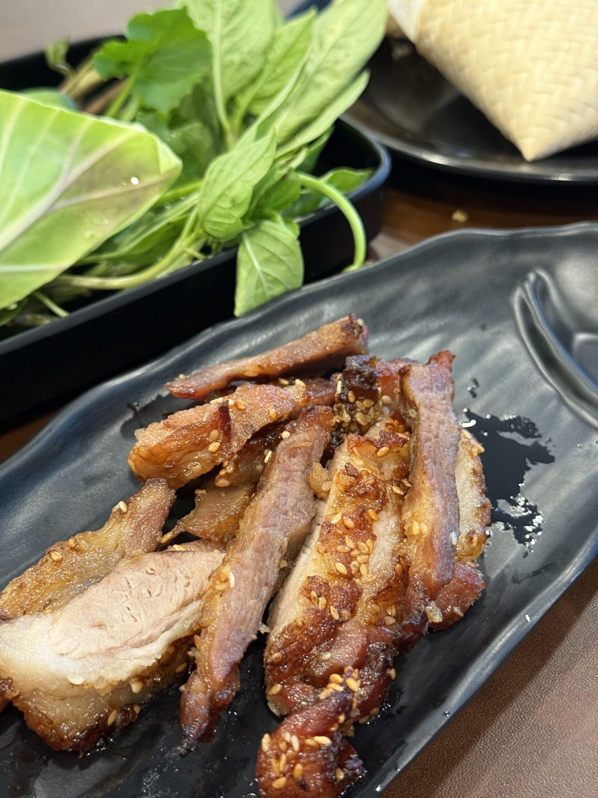 หมูสามชั้นย่างโรยงาขาว เสิร์ฟบนจานสีดำพร้อมผักสดและข้าวเหนียว