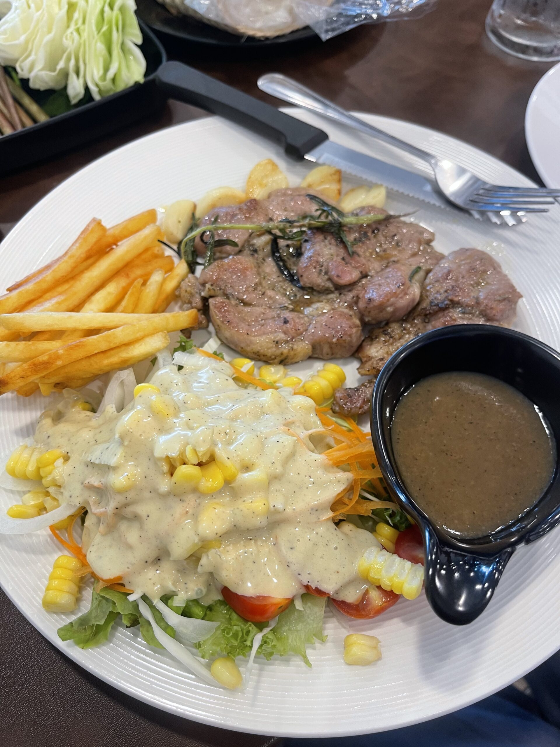 จานสเต็กหมูย่างสมุนไพรโรสแมรี่ เสิร์ฟพร้อมเฟรนช์ฟรายส์ สลัดผักราดซอสครีม ข้าวโพด และน้ำซอสพริกไทยดำในถ้วยดำ
