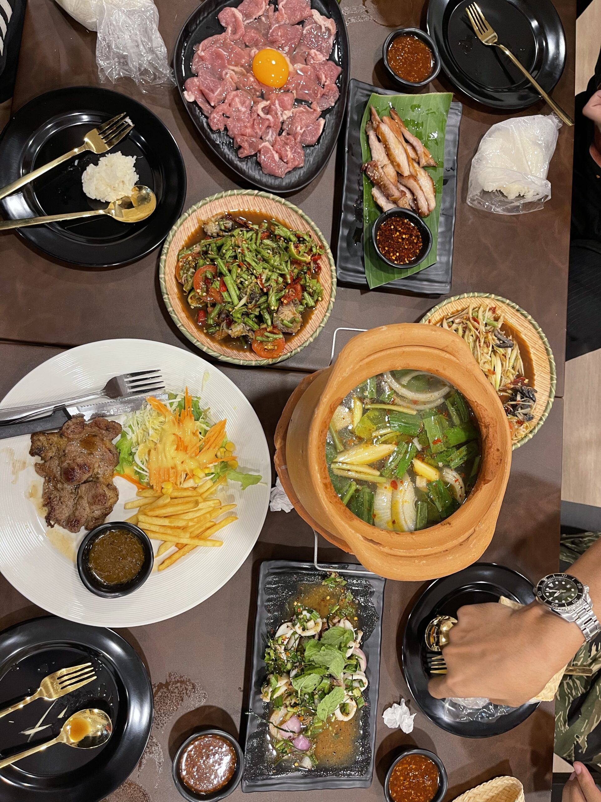 โต๊ะอาหารมองจากด้านบน มีเนื้อดิบกับไข่แดง ผัดพริกแกง ส้มตำ สเต็กเนื้อพร้อมเฟรนช์ฟรายส์ ต้มยำหม้อดิน ยำทะเล คอหมูย่าง และข้าวเหนียว