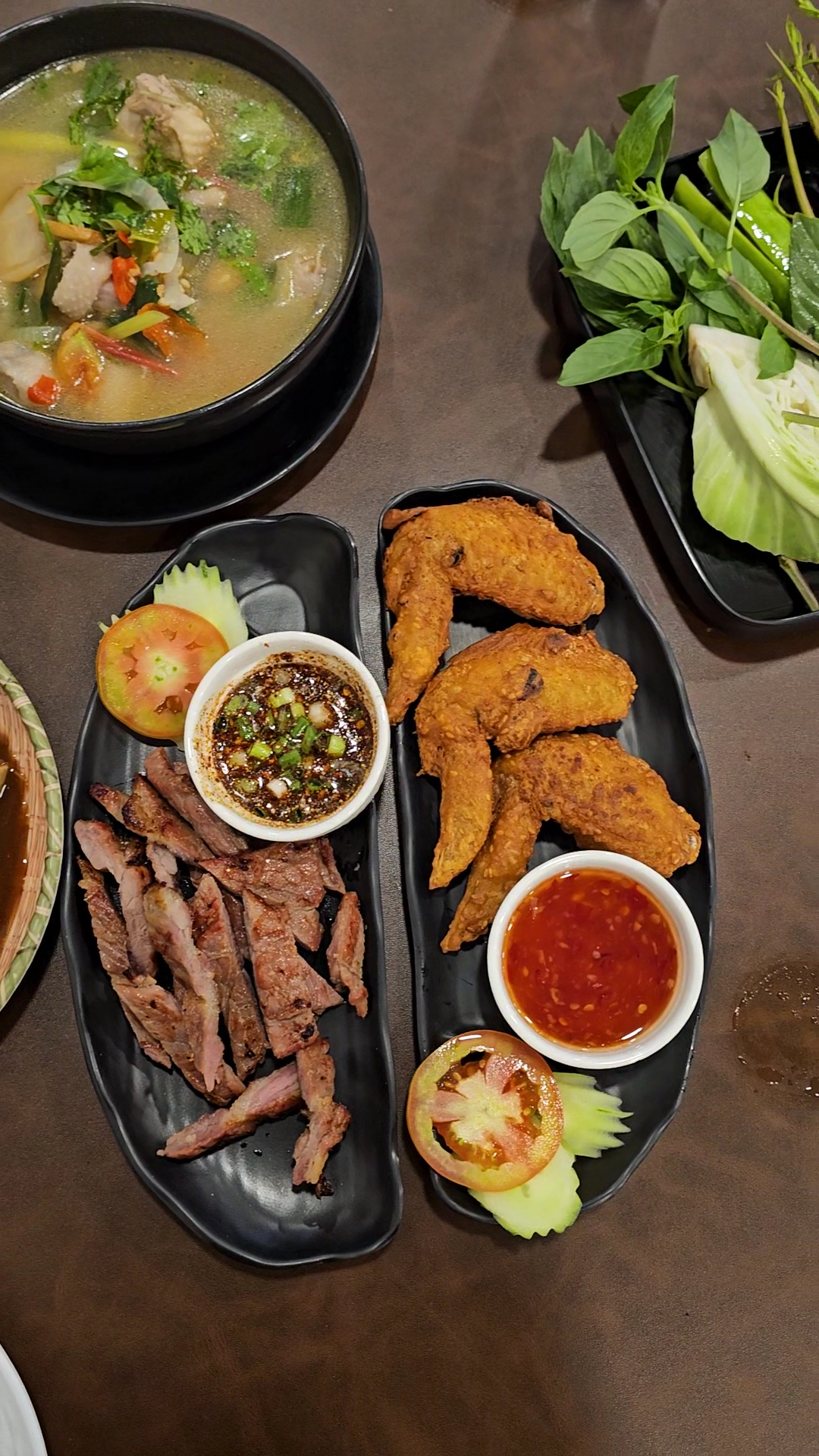 อาหารไทยบนโต๊ะ ประกอบด้วยเนื้อย่างหั่นชิ้นพร้อมน้ำจิ้มแจ่ว ปีกไก่ทอดพร้อมซอสพริก ต้มแซ่บในหม้อดำ และผักสดเคียง