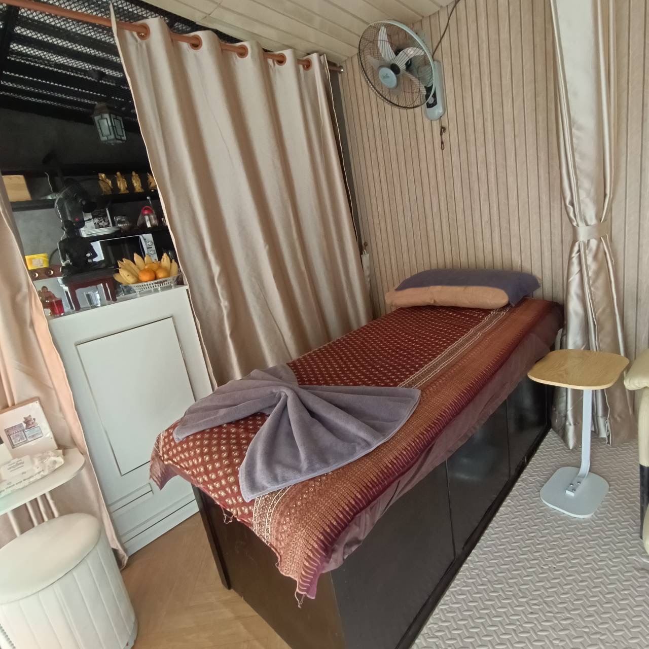 ห้องนวดไทยพร้อมเตียงนวดปูผ้าลายไทยสีแดง มีพัดลมติดผนังและม่านกั้น