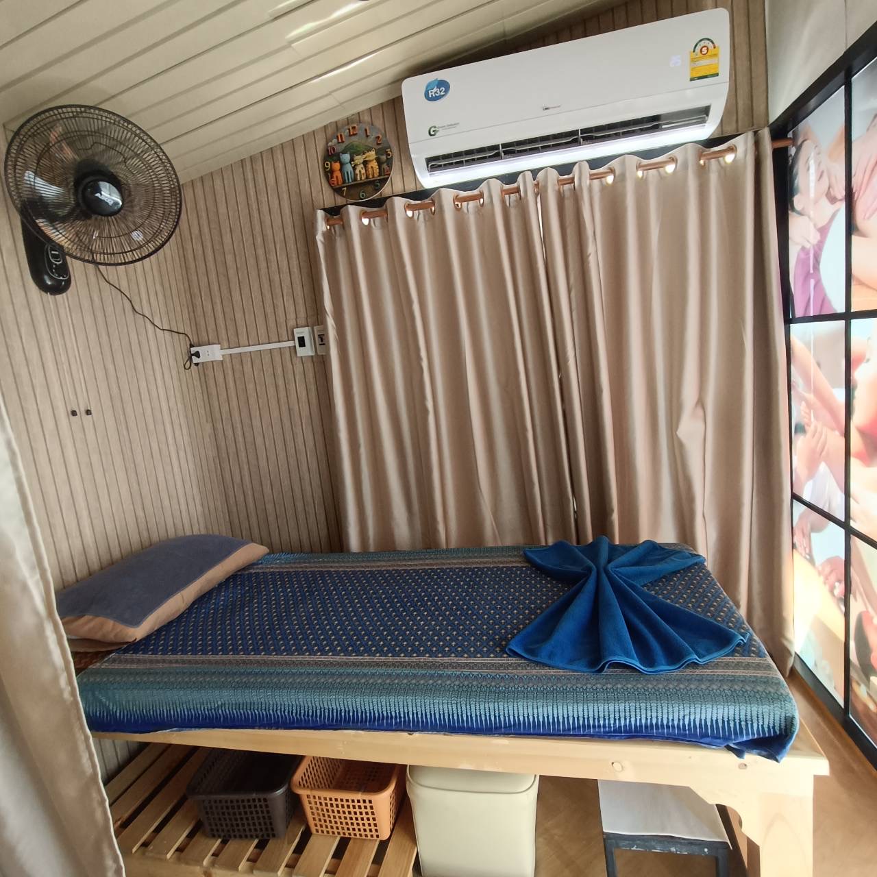 ห้องนวดไทยพร้อมเตียงนวดผ้าลายไทยสีน้ำเงิน มีแอร์และพัดลมติดผนัง พร้อมผ้าม้วนตกแต่งรูปพัด