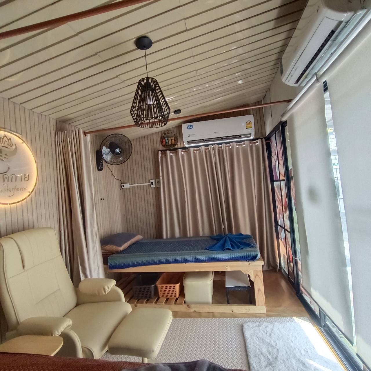 ห้องนวดไทยตกแต่งอบอุ่นมีเตียงนวด เก้าอี้นวด ผ้าม่านสีน้ำตาล พัดลมติดผนัง และแอร์
