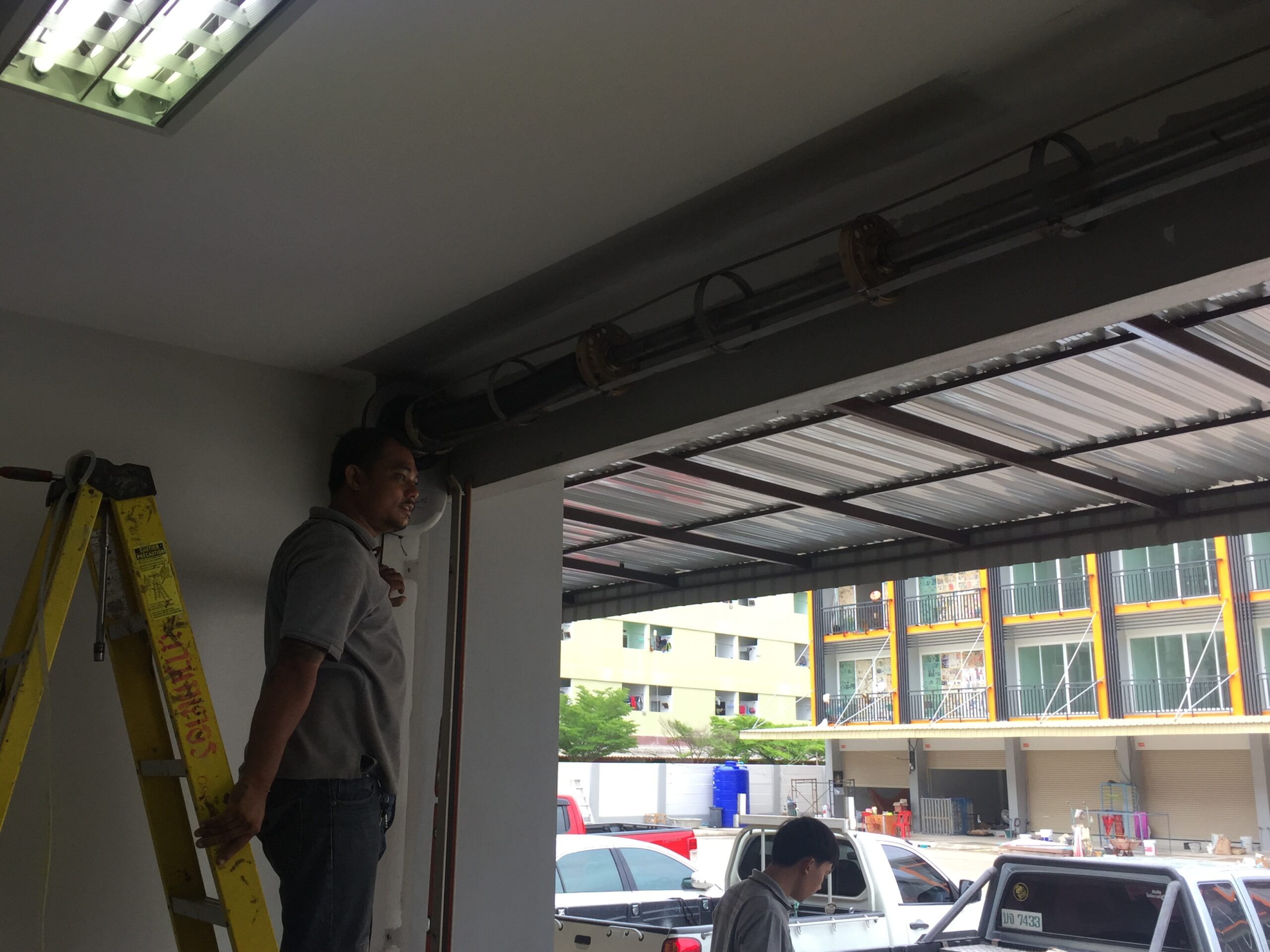 ช่างกำลังติดตั้งประตูม้วนเหล็กที่หน้าร้าน โดยยืนบนบันไดสีเหลืองตรวจสอบรางประตูด้านบน