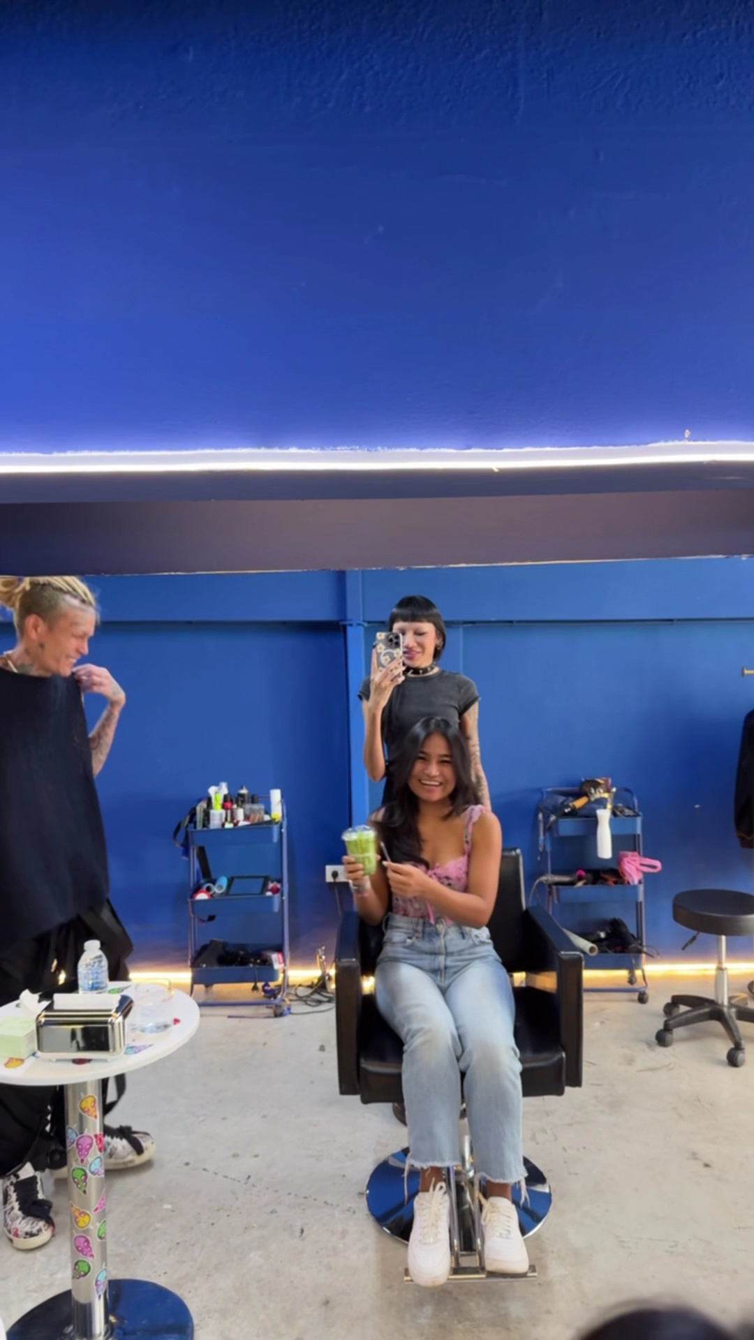 ลูกค้าสาวนั่งยิ้มถือเครื่องดื่มสีเขียวบนเก้าอี้ตัดผมในร้านทำผมสีน้ำเงิน ขณะช่างทำผมถ่ายรูปผ่านกระจก