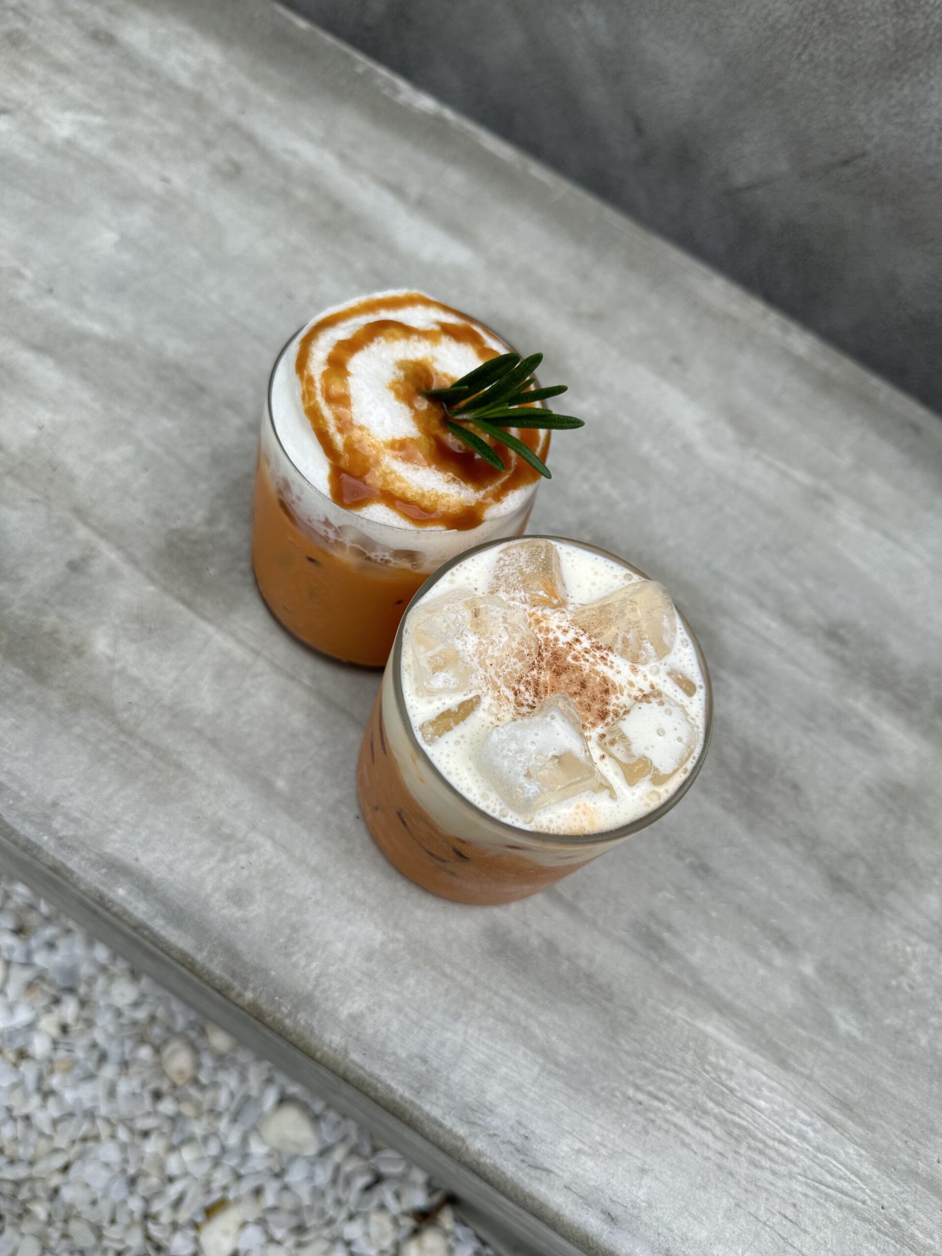 เครื่องดื่มชาไทยเย็นสองแก้วบนโต๊ะปูน ตกแต่งด้วยโฟมนม ซอสคาราเมล ใบโรสแมรี่ และผงอบเชย