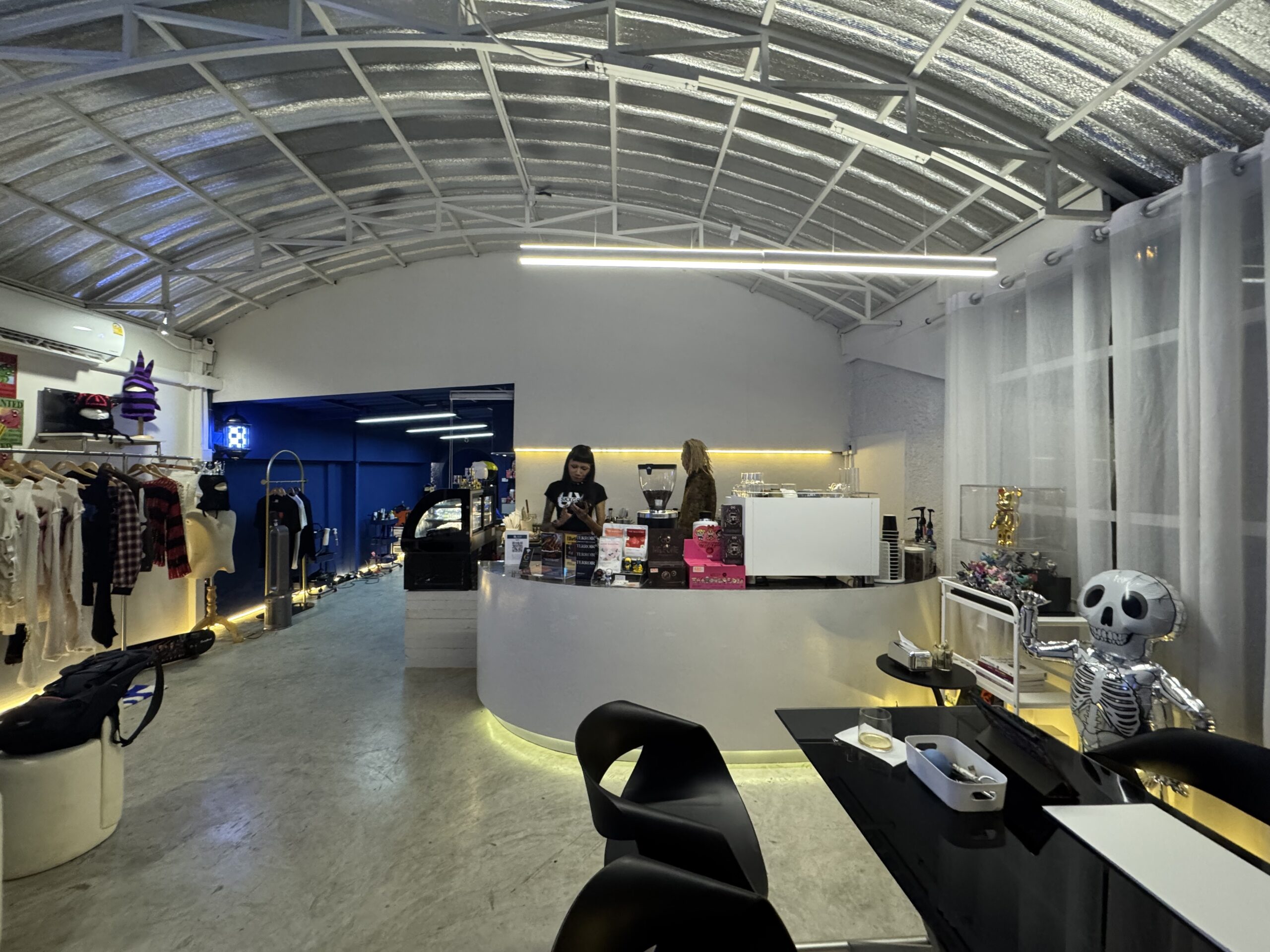 ภายในร้านกาแฟสไตล์อินดัสเทรียลหลังคาโค้งเหล็ก มีเคาน์เตอร์สีขาวโค้งพร้อมเครื่องชงกาแฟ ตกแต่งด้วยตุ๊กตาโครงกระดูกสีเงินและราวแขวนเสื้อผ้า