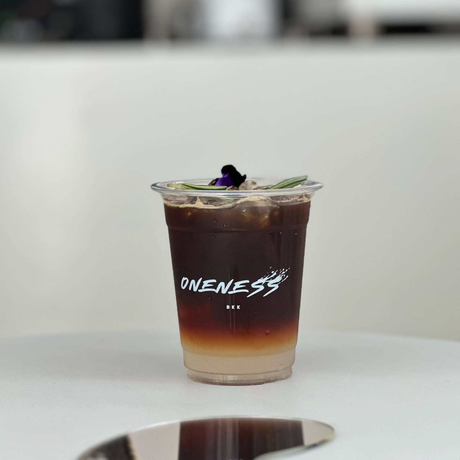 เครื่องดื่มกาแฟเย็นแบบเลเยอร์ในแก้วพลาสติกแบรนด์ Oneness BKK ตกแต่งด้วยดอกอัญชันและโรสแมรี่ วางบนโต๊ะสีขาว