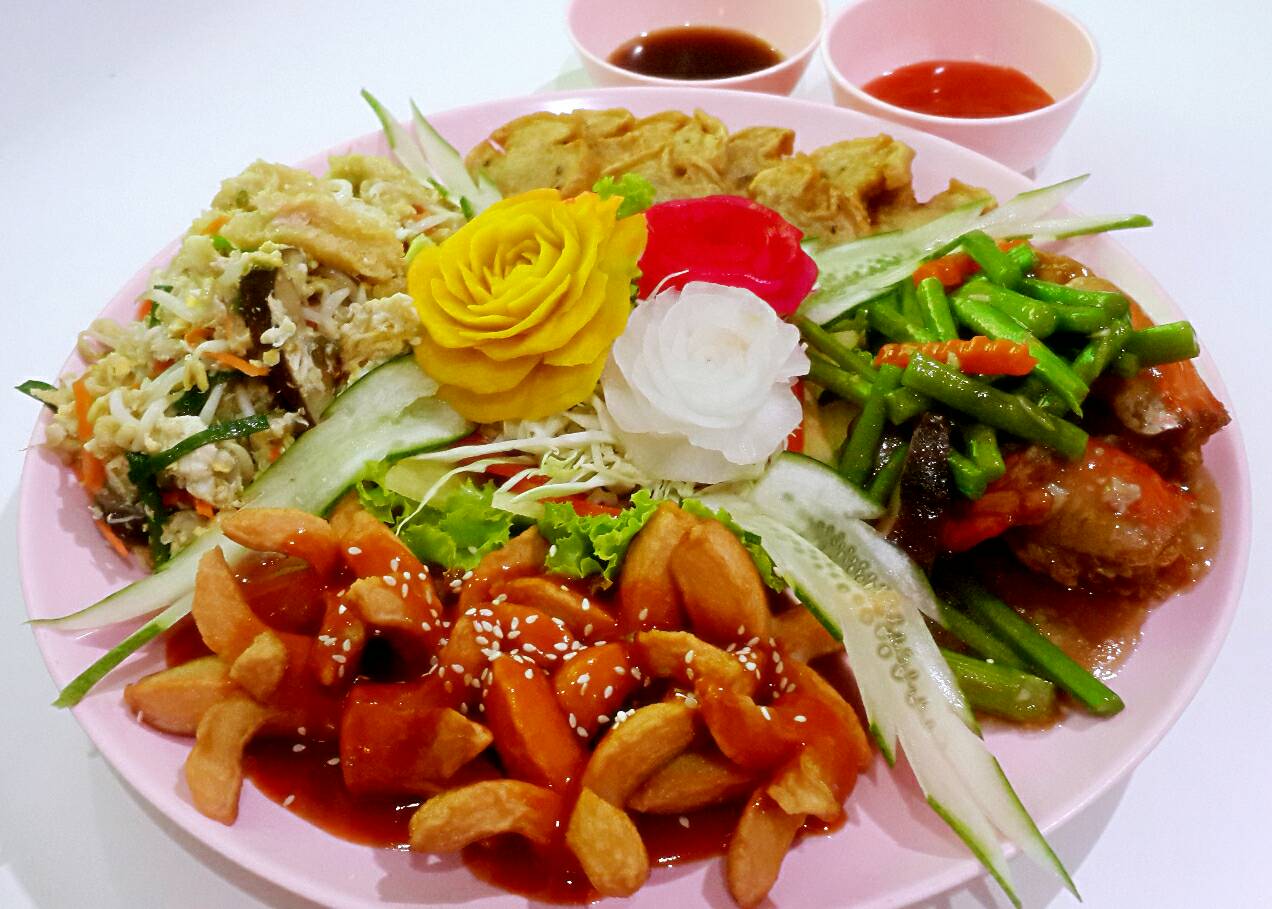 จานอาหารเจรวมบนจานสีชมพู มีผัดผักรวม เต้าหู้ทอด ผัดเปรี้ยวหวาน และผัดถั่วฝักยาว ตกแต่งด้วยดอกไม้แกะสลักจากผักสวยงาม พร้อมน้ำจิ้ม 2 ถ้วย