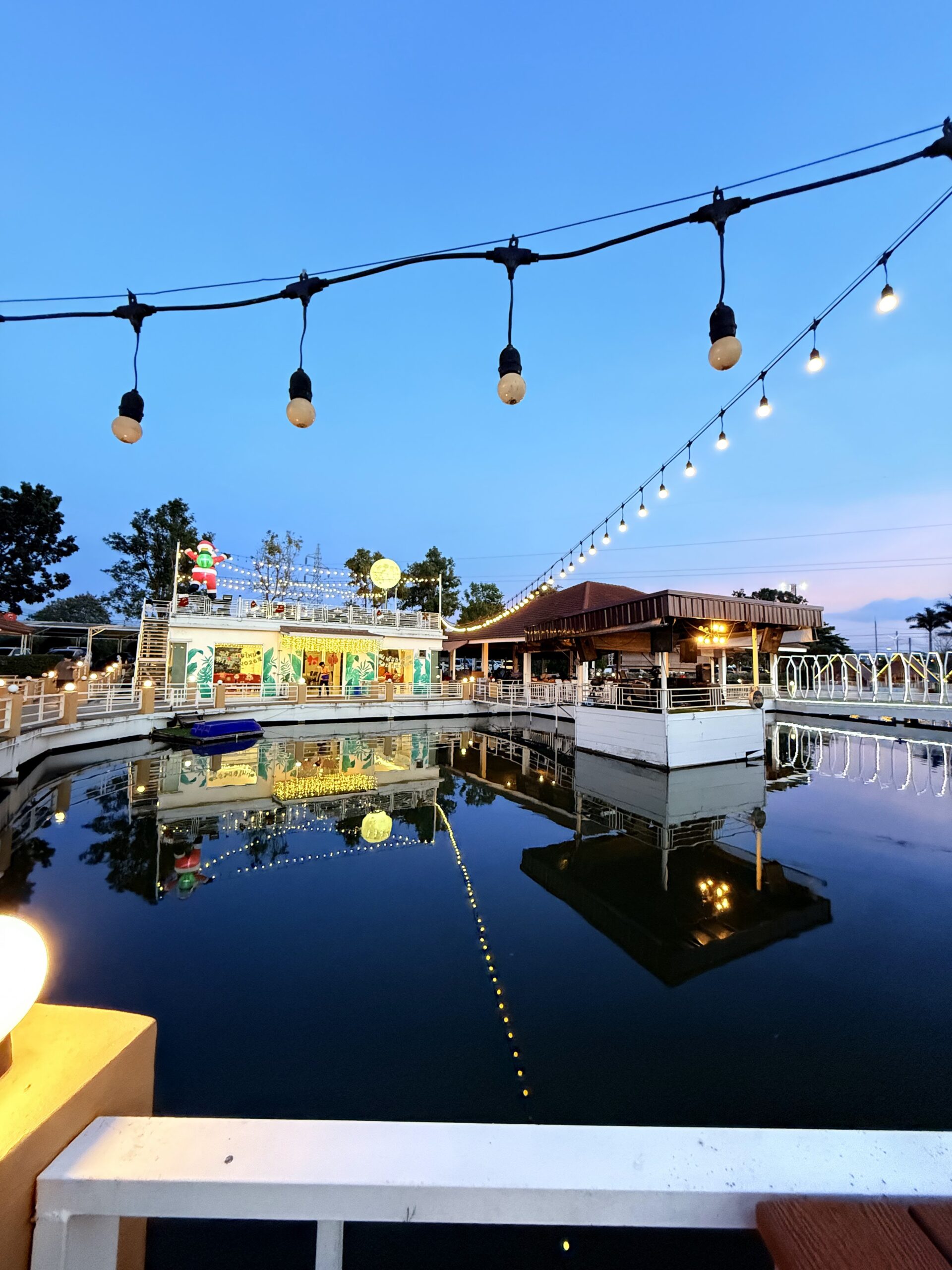 ร้านอาหารริมน้ำยามพลบค่ำ ตกแต่งด้วยไฟสายประดับและไฟหลากสี มีสระน้ำสะท้อนแสงไฟสวยงาม พร้อมตุ๊กตาซานตาคลอสบนหลังคา