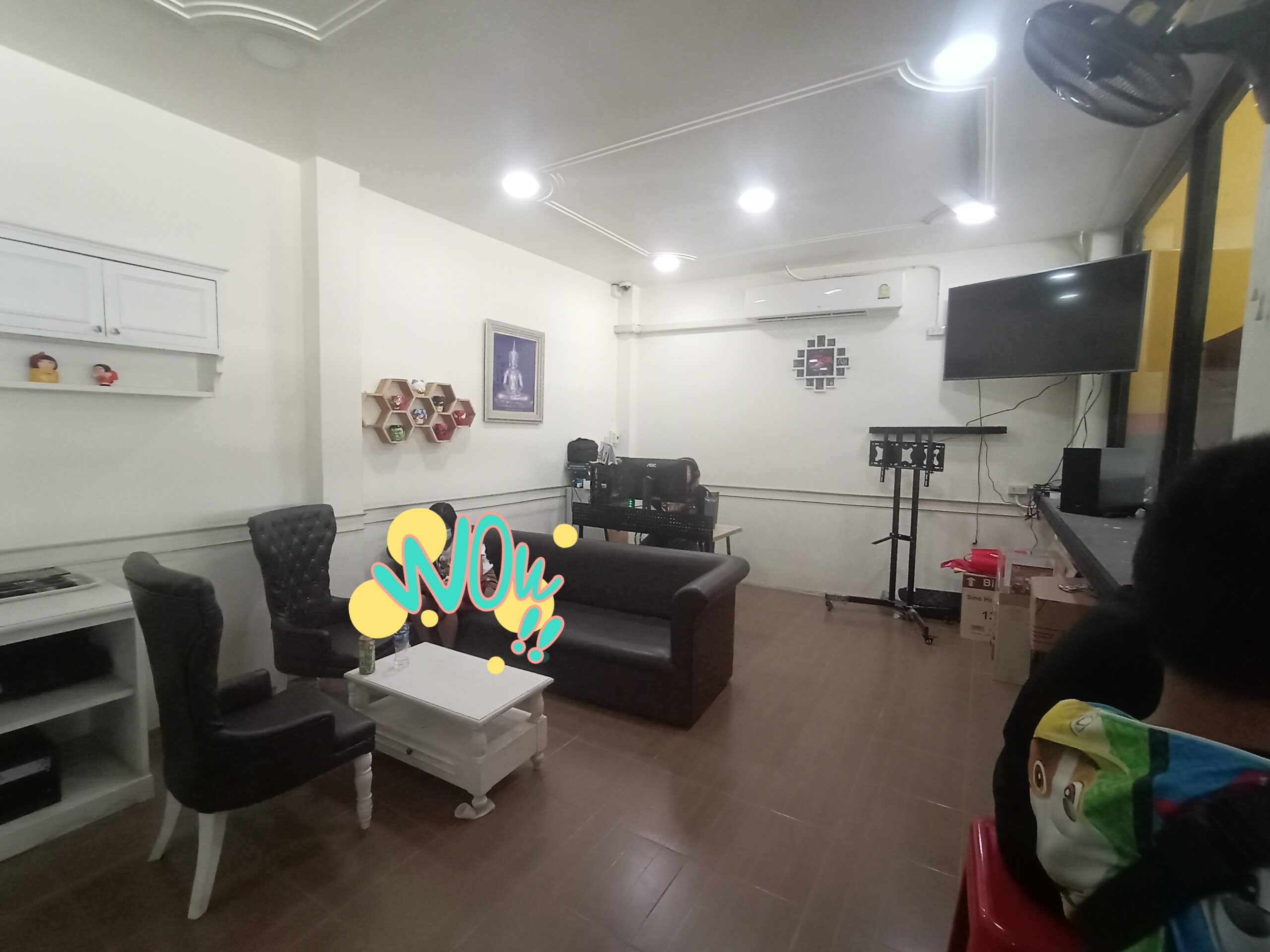 ห้องรับรองภายในร้านอาหารพร้อมโซฟาหนังสีดำ เก้าอี้หลุยส์ โต๊ะกลางสีขาว ทีวีจอแบน และแอร์ ตกแต่งด้วยชั้นวางรูปทรงรังผึ้งและรูปภาพบนผนัง