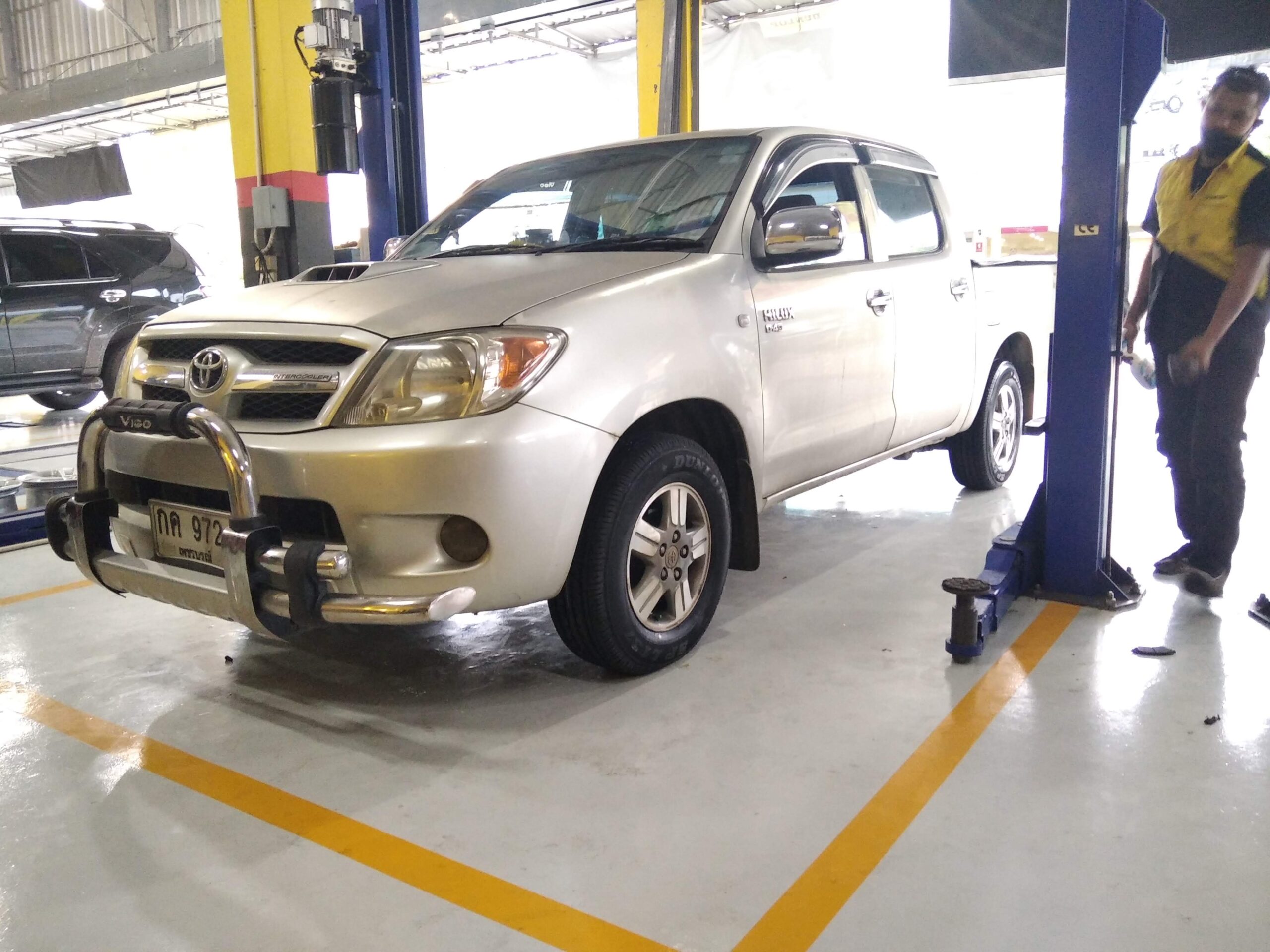 รถกระบะโตโยต้า ไฮลักซ์ วีโก้ สีเงิน จอดอยู่ในศูนย์บริการซ่อมรถยนต์ มีช่างยืนอยู่ข้างรถ