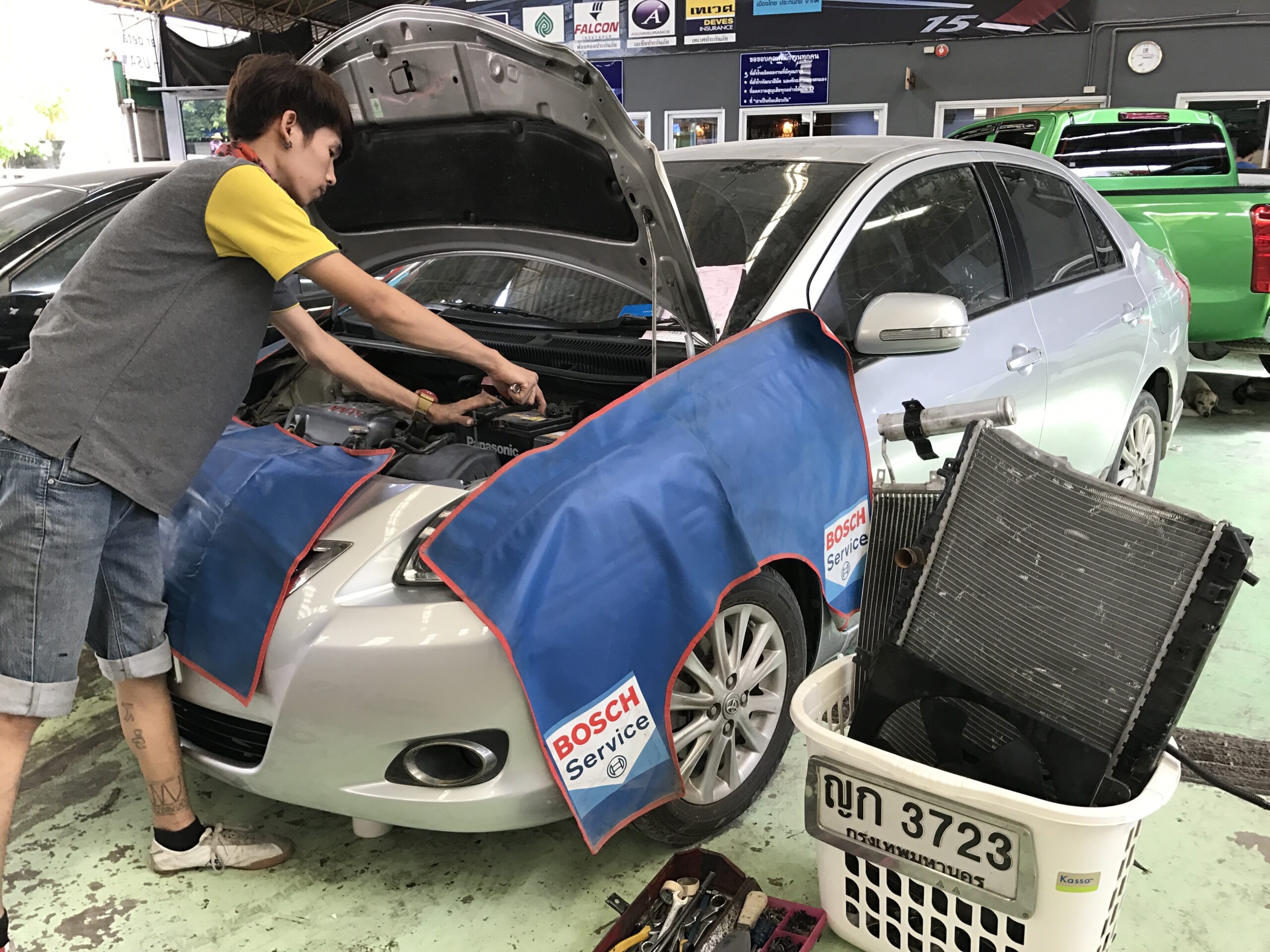 ช่างซ่อมรถยนต์กำลังซ่อมเครื่องยนต์รถสีเงินที่ศูนย์บริการ Bosch Service พร้อมผ้าคลุมป้องกันสีน้ำเงิน