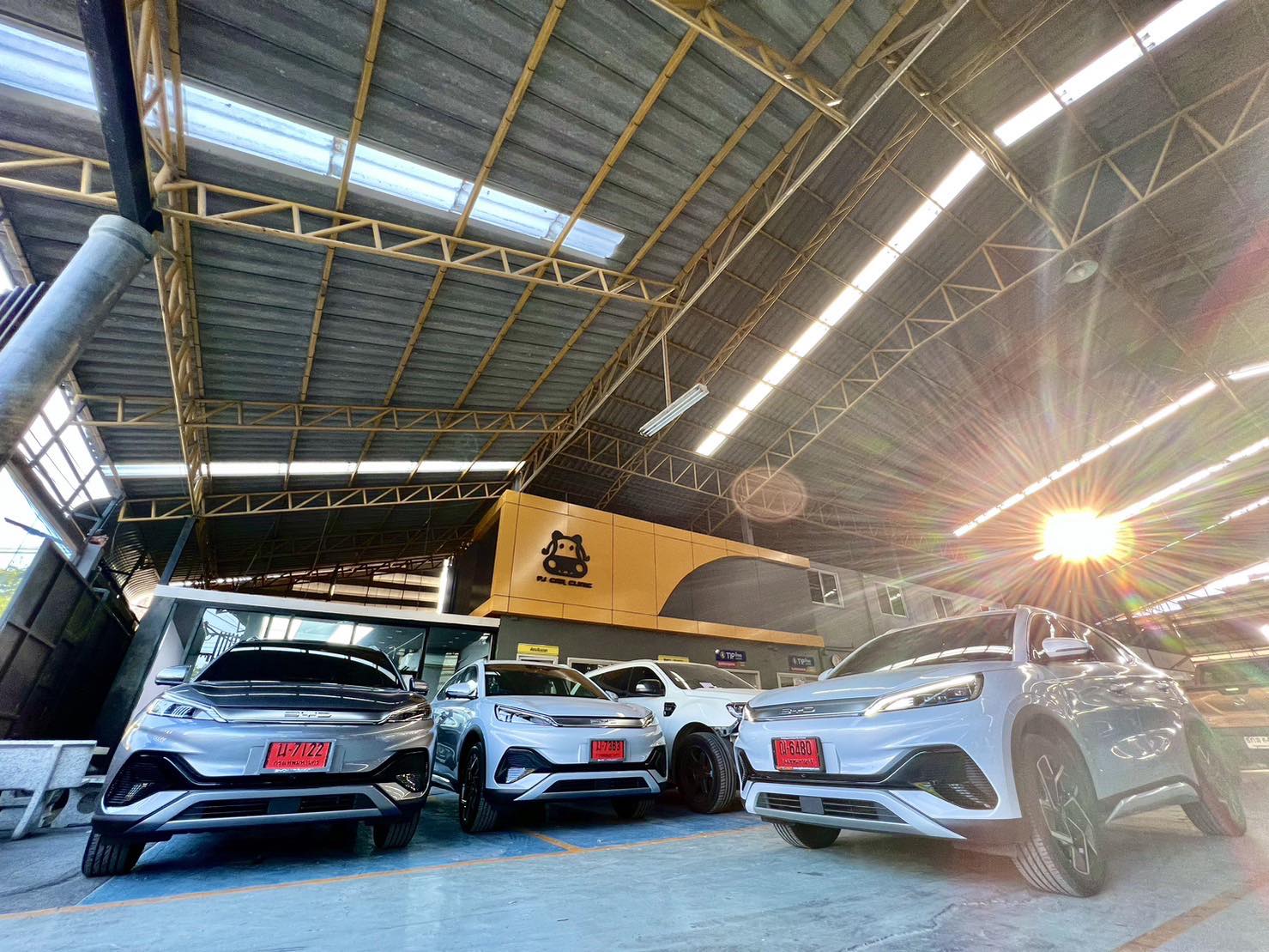 รถยนต์ BYD สีขาวและสีเทาจอดเรียงกันในโชว์รูมโกดังที่มีโครงหลังคาเหล็กและแสงแดดส่องเข้ามา
