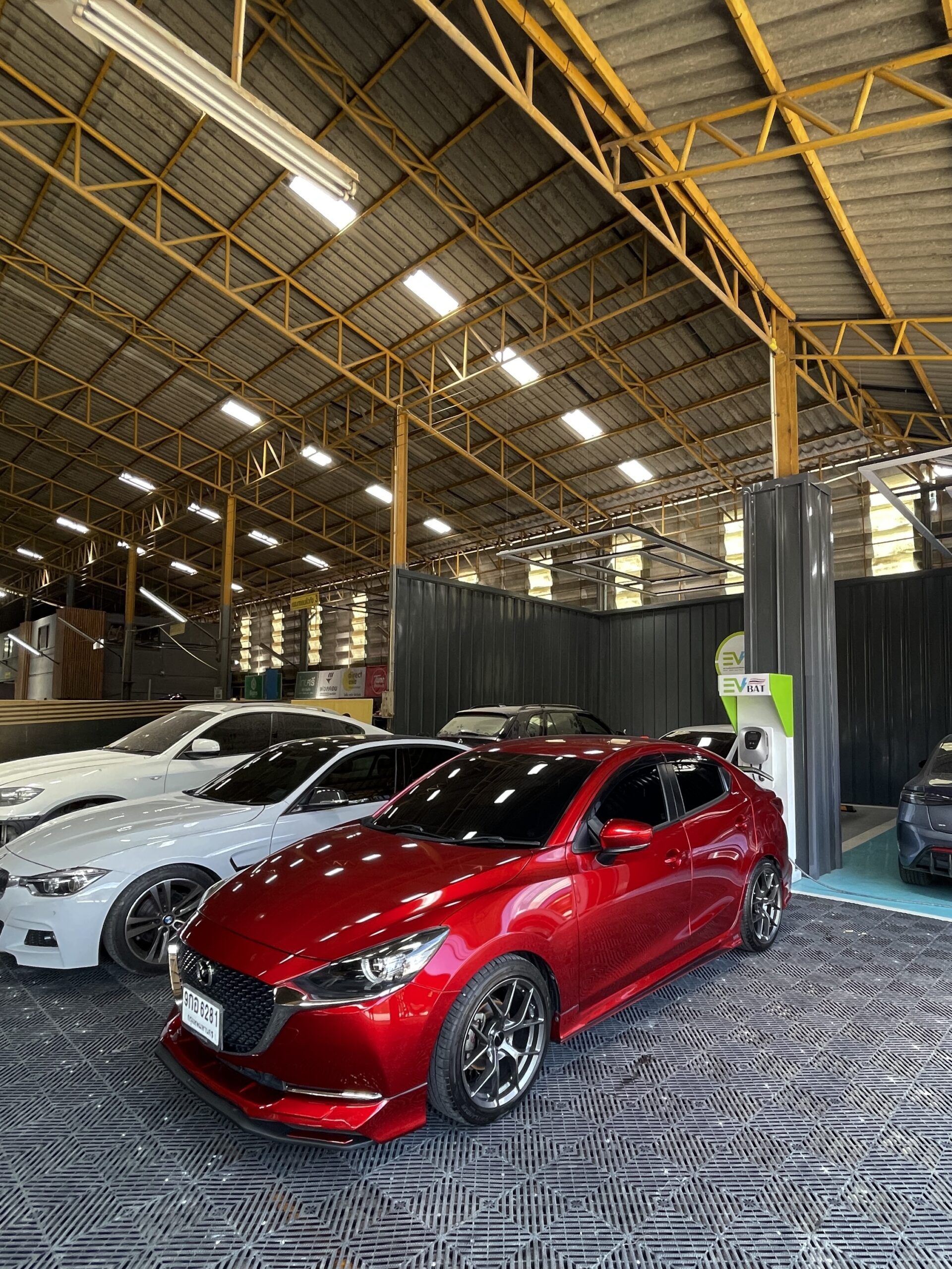 รถยนต์มาสด้าสีแดงจอดในโกดังที่มีโครงหลังคาเหล็กสีเหลือง พร้อมรถยนต์คันอื่นๆ จอดอยู่ด้านหลัง