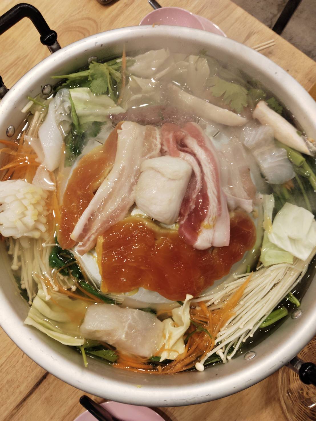 หม้อสุกี้ร้อนๆ ที่เต็มไปด้วยเนื้อหมูสไลด์ เห็ดเข็มทอง ผักกาดขาว ผักชี แครอทซอย และน้ำซุปใส บนโต๊ะไม้ในร้านอาหาร