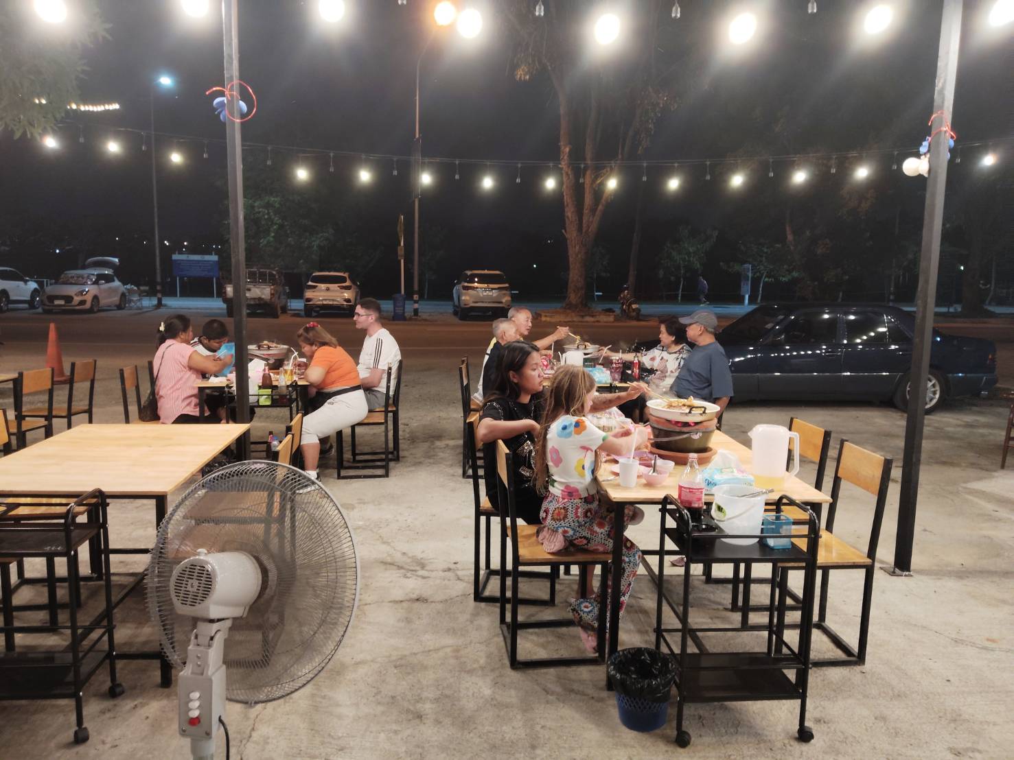ร้านอาหารกลางแจ้งยามค่ำคืนมีลูกค้านั่งทานอาหารหลายโต๊ะ ตกแต่งด้วยไฟสายสวยงาม มีพัดลมตั้งพื้นและที่จอดรถด้านหลัง